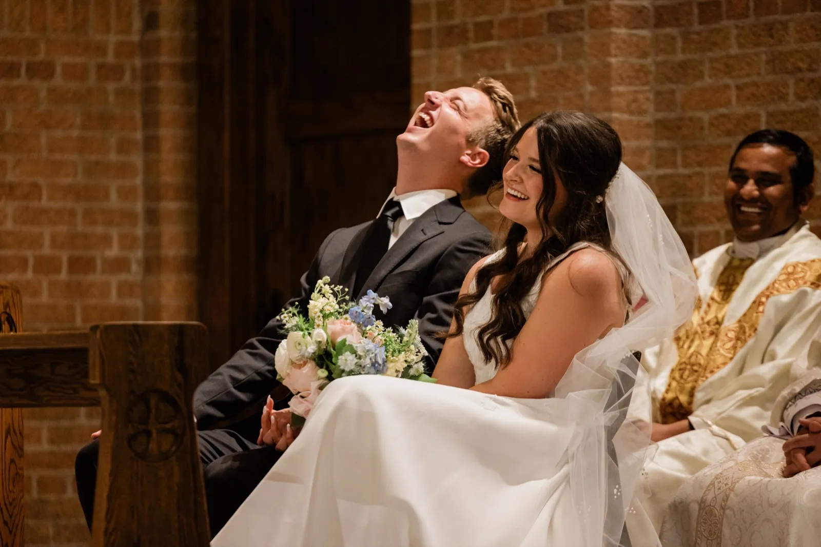 The groom throws his head back laughing beside the bride during their nuptial Mass, the priest seated beside them laughing too — Tim Larsen Photography, Brainerd Lakes MN