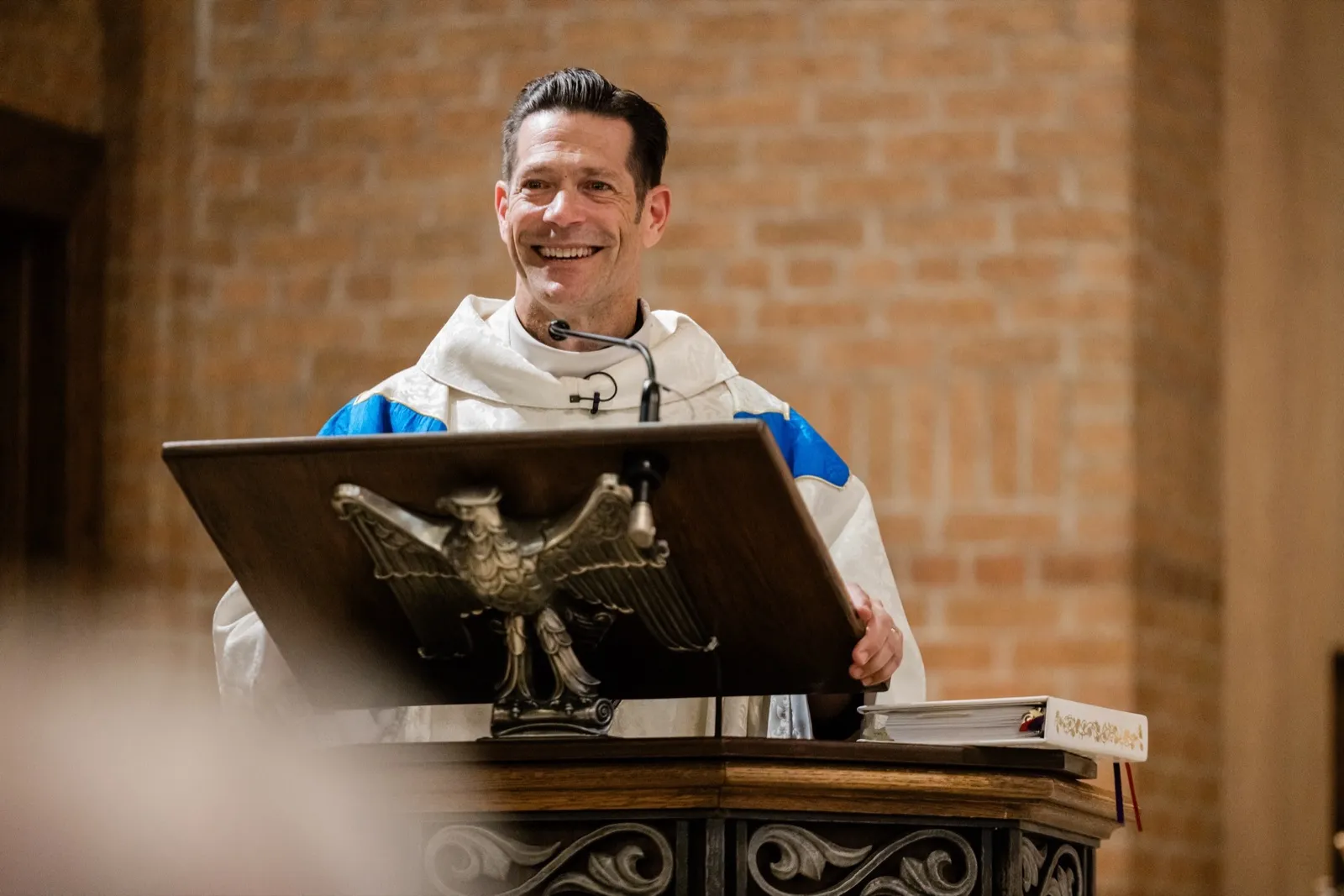A priest laughs while delivering the homily from a brass eagle lectern in a brick-walled Catholic church — Tim Larsen Photography, Brainerd Lakes MN