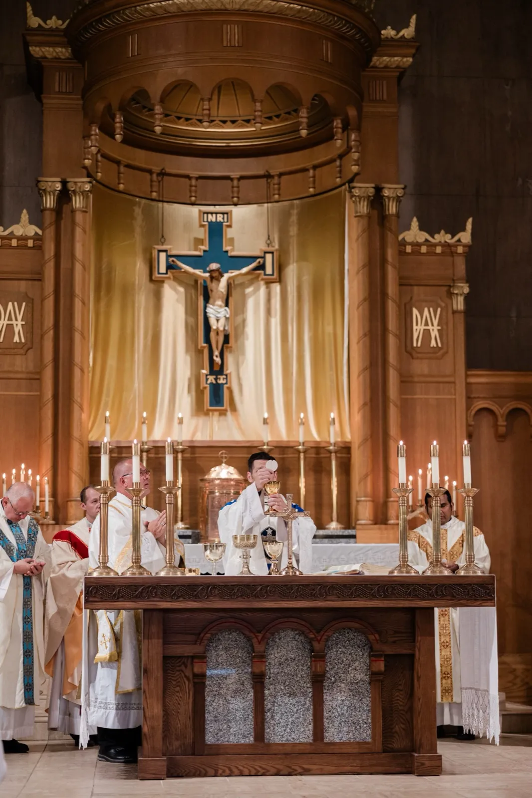 A wide view of the church sanctuary shows a priest elevating the host beneath a large crucifix, flanked by candles and concelebrating clergy — Tim Larsen Photography, Brainerd Lakes MN