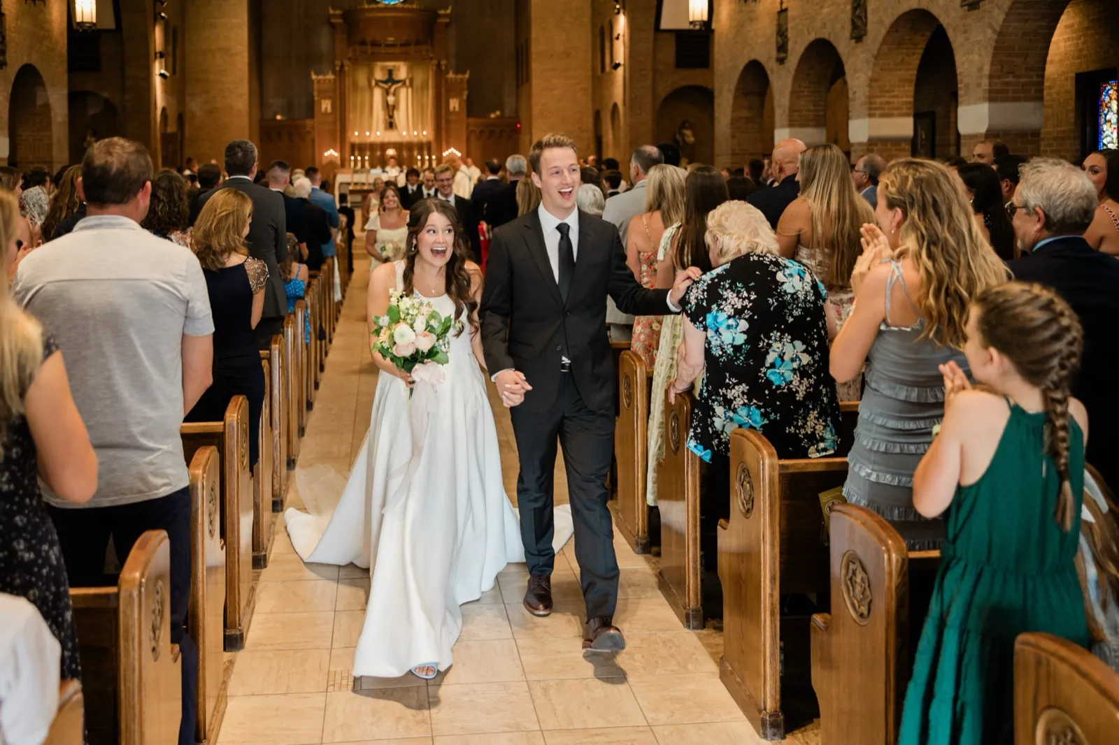The newly married couple laugh with joy as they walk back down the aisle, the bride holding her bouquet while guests applaud from the pews — Tim Larsen Photography, Brainerd Lakes MN