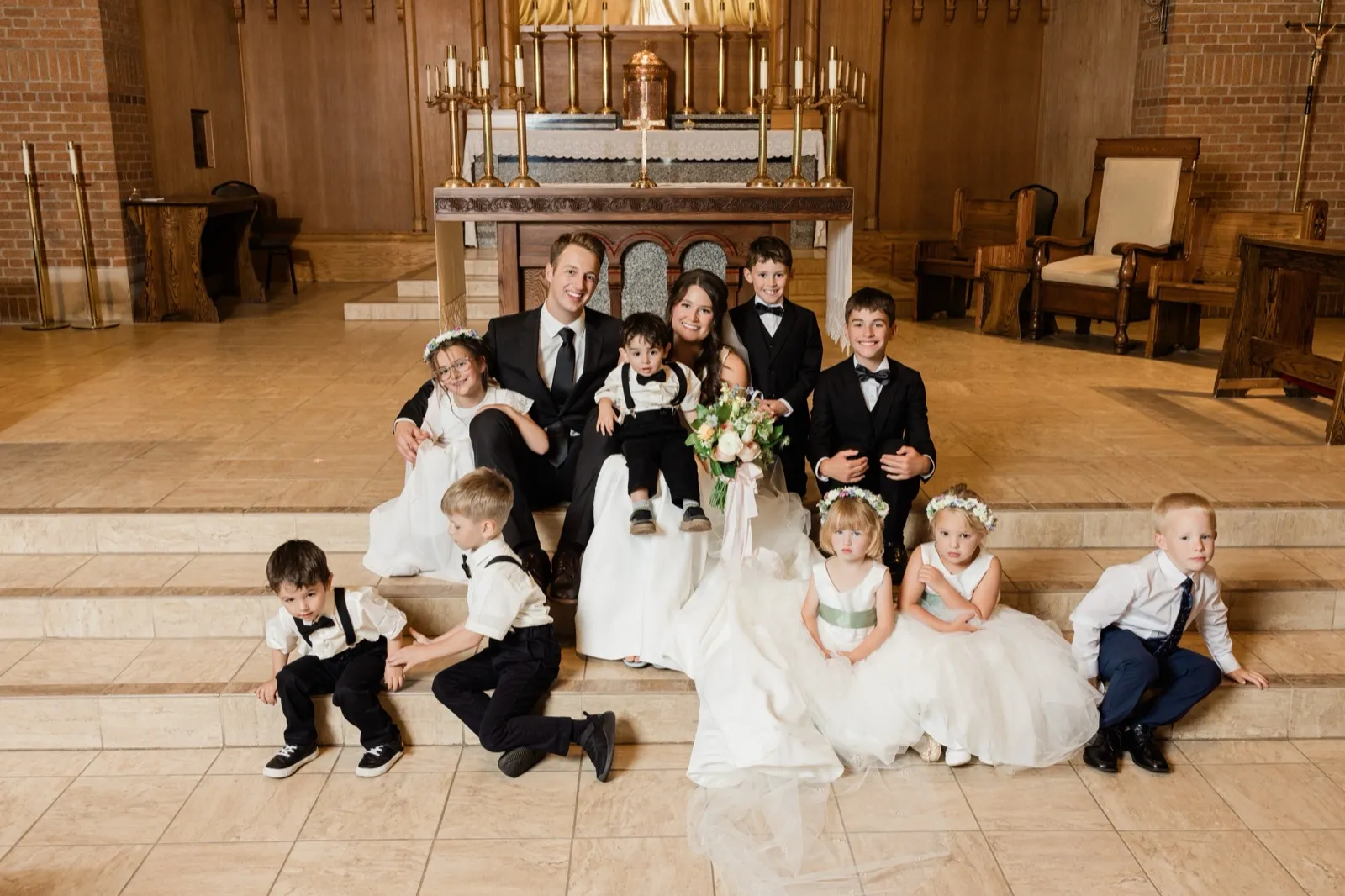 The bride and groom sit on the church altar steps surrounded by all the young flower girls and ring bearers in formal attire — Tim Larsen Photography, Brainerd Lakes MN