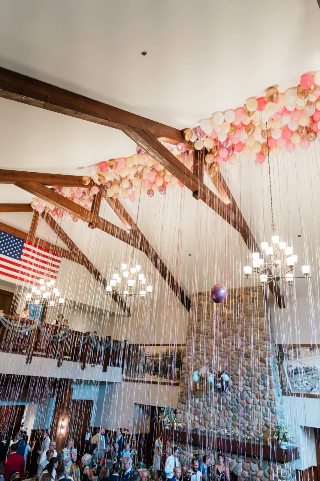 A cascade of pastel balloons and streamers falls from the timber beams past the great room's tall fieldstone chimney to the cocktail-hour guests below — Tim Larsen Photography, Brainerd Lakes MN
