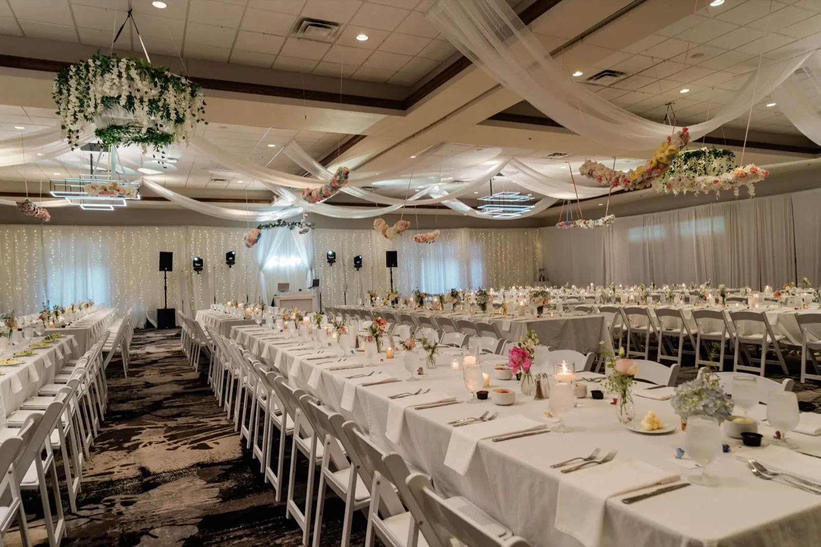 Long banquet tables set with white linens, candles, and floral centerpieces fill the reception ballroom beneath draped ceiling fabric and hanging floral hoops — Tim Larsen Photography, Brainerd Lakes MN