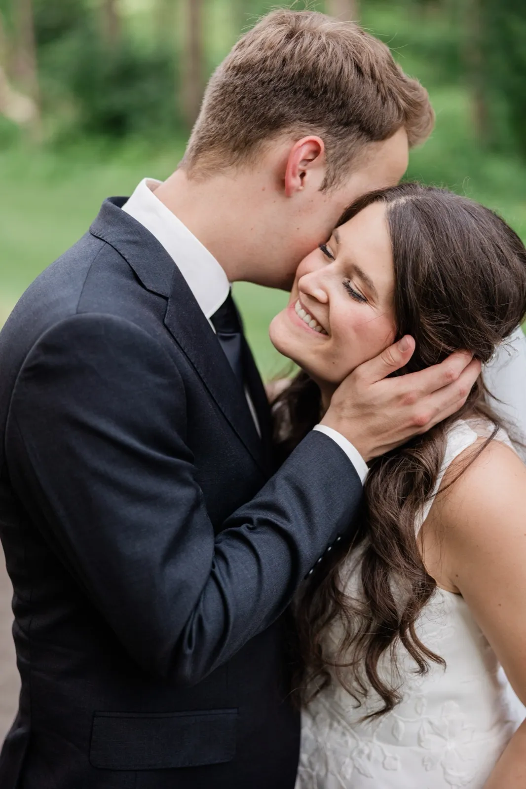 The groom leans in to nuzzle the smiling bride's temple, his hand cradling her hair, against a backdrop of soft green woods — Tim Larsen Photography, Brainerd Lakes MN