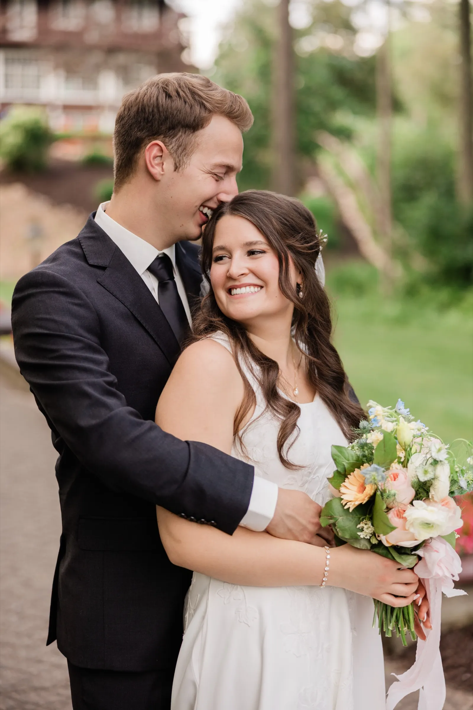 The groom laughs into the bride's hair as she smiles brightly at the camera, holding her pastel bouquet close, the lodge grounds soft behind them — Tim Larsen Photography, Brainerd Lakes MN