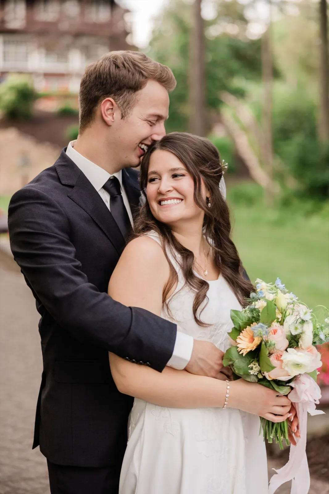 The groom laughs into the bride's hair as she smiles brightly at the camera, holding her pastel bouquet close, the lodge grounds soft behind them — Tim Larsen Photography, Brainerd Lakes MN