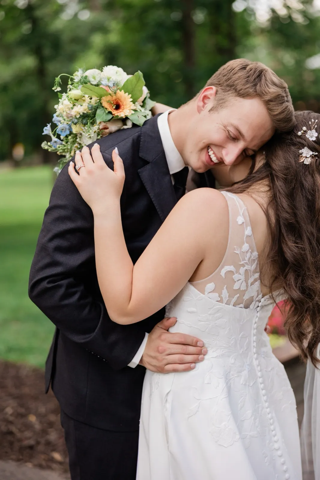 The groom laughs with his eyes closed as the bride leans into his neck, her ringed hand on his lapel, surrounded by green trees — Tim Larsen Photography, Brainerd Lakes MN