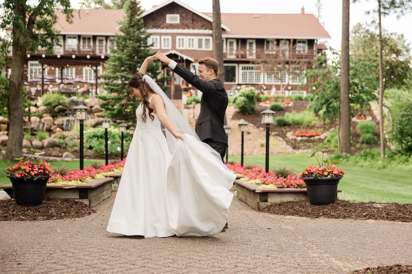The groom spins the bride under his arm, her gown and veil swinging out, with the lodge and garden beds behind them — Tim Larsen Photography, Brainerd Lakes MN