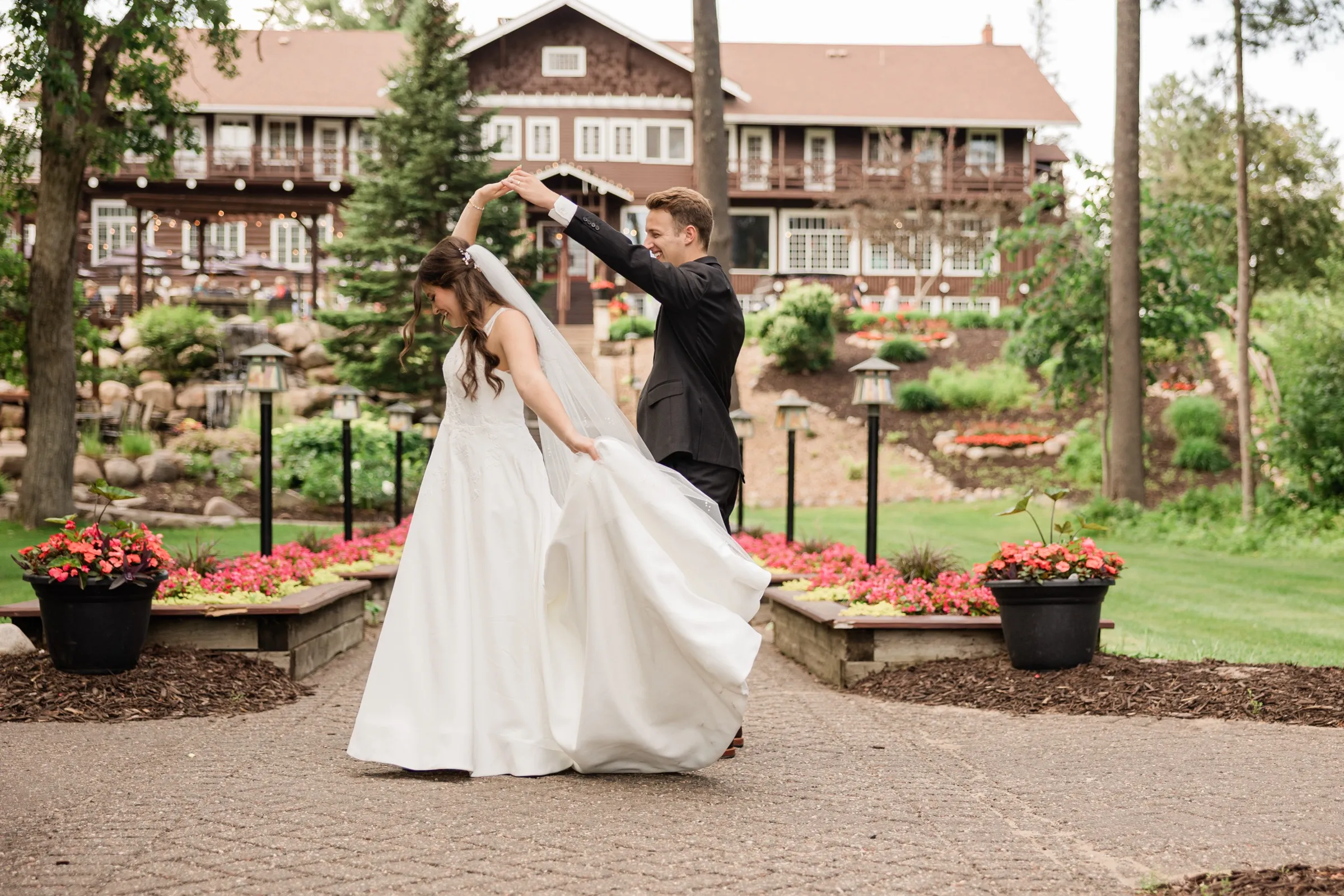 The groom spins the bride under his arm, her gown and veil swinging out, with the lodge and garden beds behind them — Tim Larsen Photography, Brainerd Lakes MN