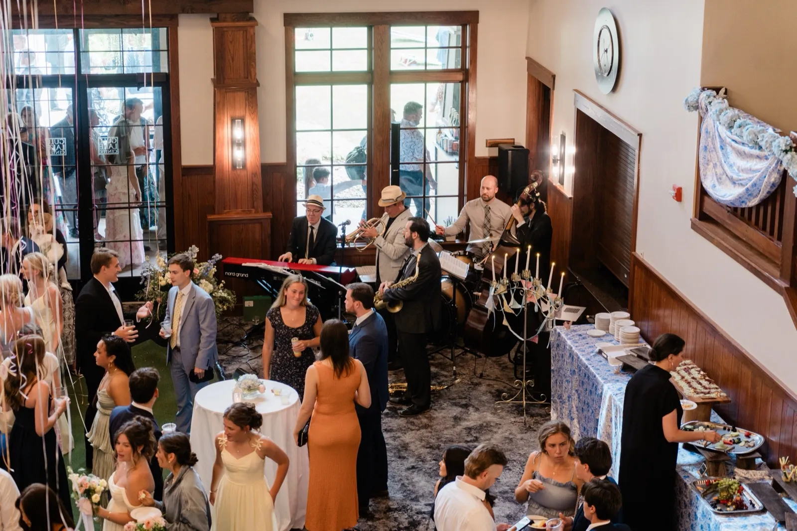 A high-angle view of wedding guests mingling with drinks as a five-piece jazz band plays under tall windows in a wood-paneled room — Tim Larsen Photography, Brainerd Lakes MN
