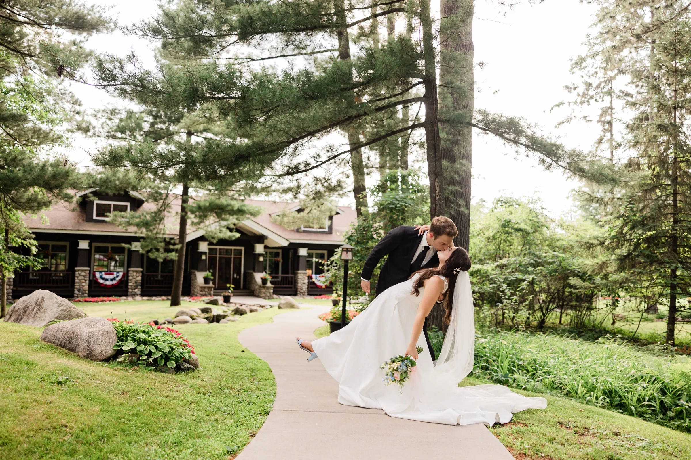 The groom dips the bride into a kiss on a curved walkway, her foot lifted, in front of a craftsman cabin draped with patriotic bunting — Tim Larsen Photography, Brainerd Lakes MN