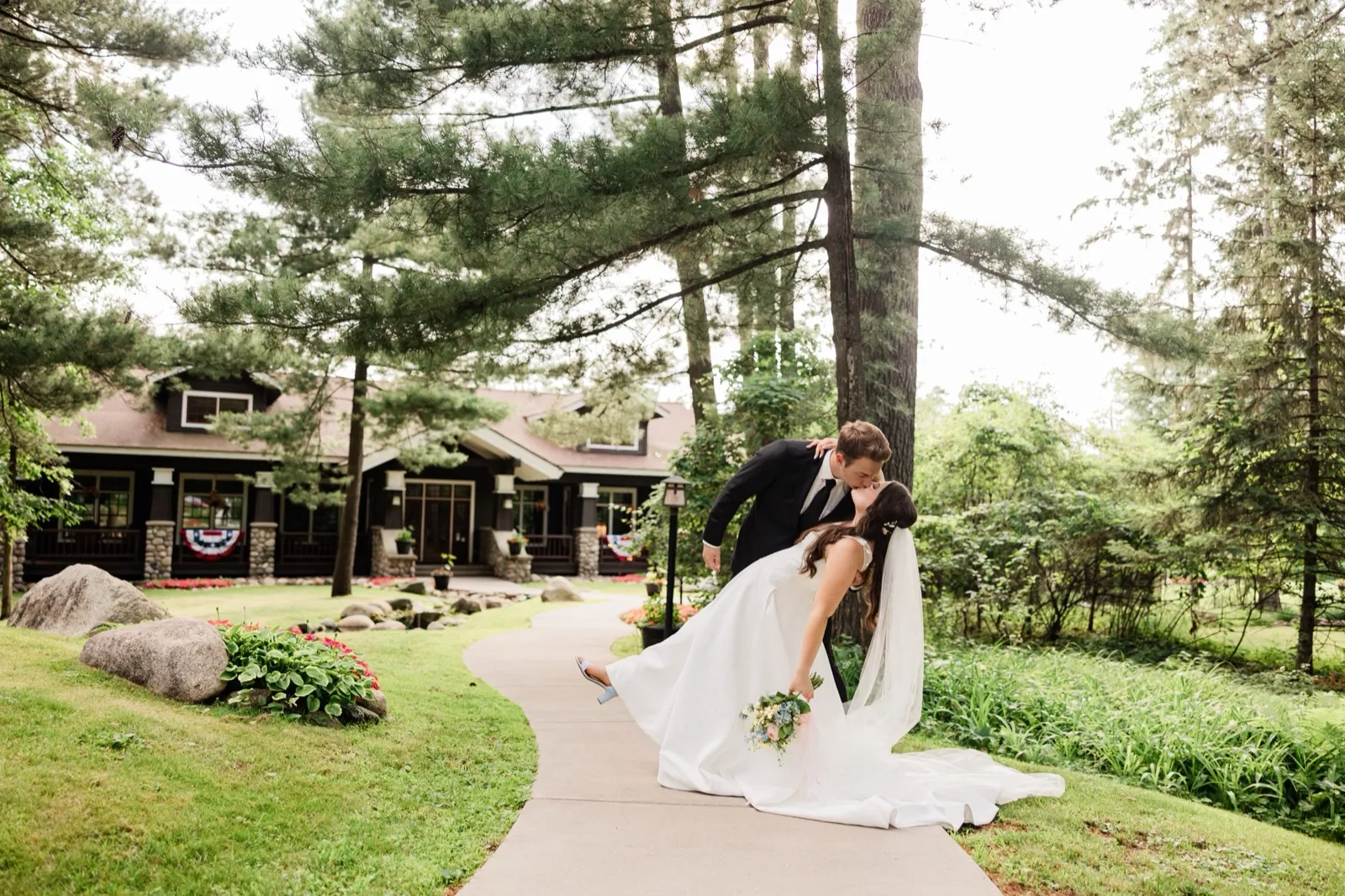 The groom dips the bride into a kiss on a curved walkway, her foot lifted, in front of a craftsman cabin draped with patriotic bunting — Tim Larsen Photography, Brainerd Lakes MN