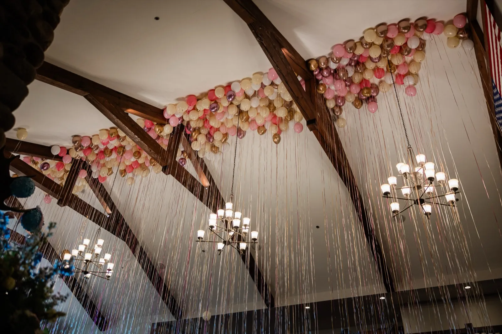 Hundreds of blush, gold, and cream balloons cover a timber-beam ceiling with ribbons cascading down between lit chandeliers, a small American flag at the edge — Tim Larsen Photography, Brainerd Lakes MN