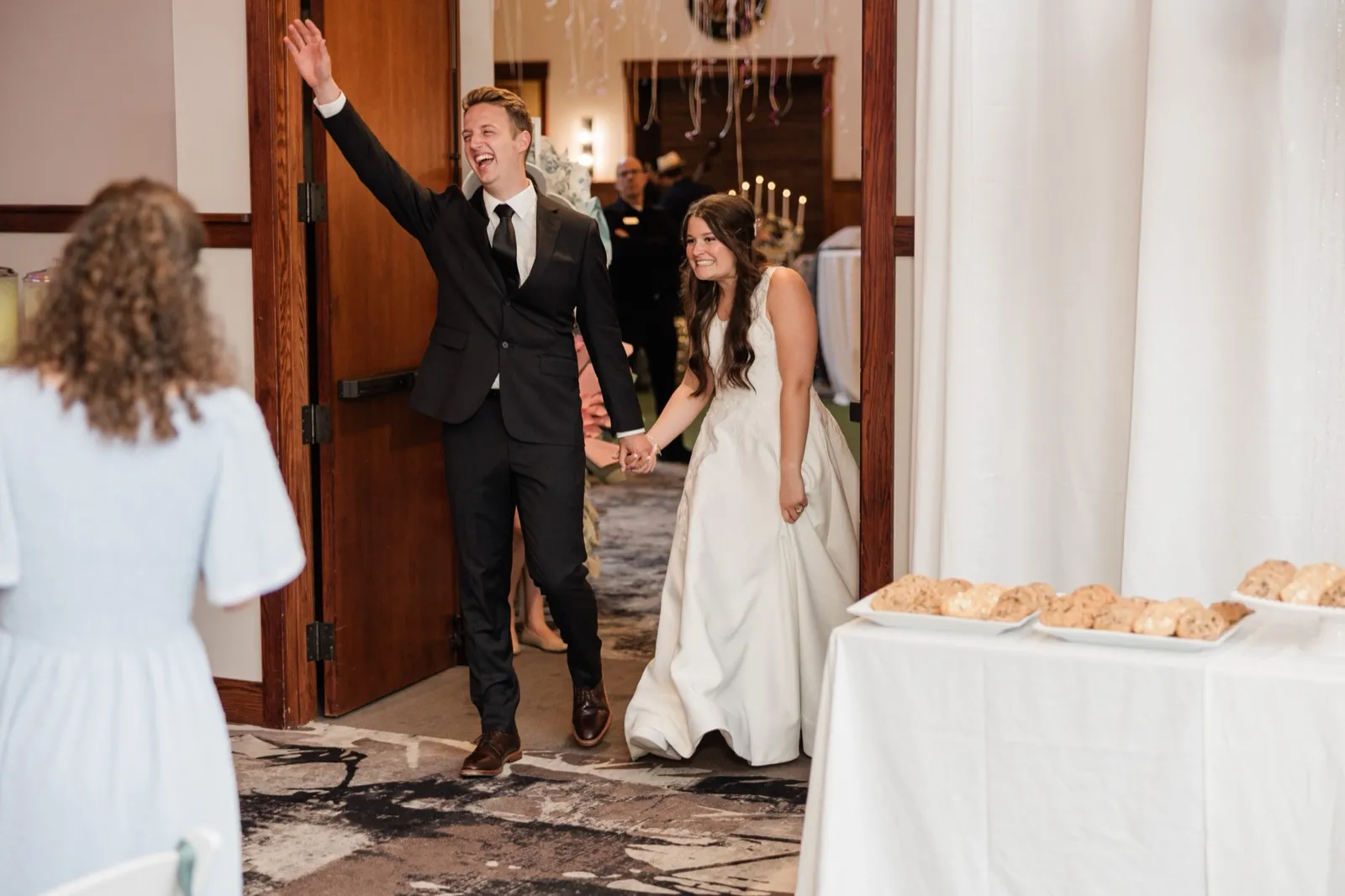 The groom throws one arm up cheering as he and the bride enter the reception hand in hand past a dessert table of cookies — Tim Larsen Photography, Brainerd Lakes MN