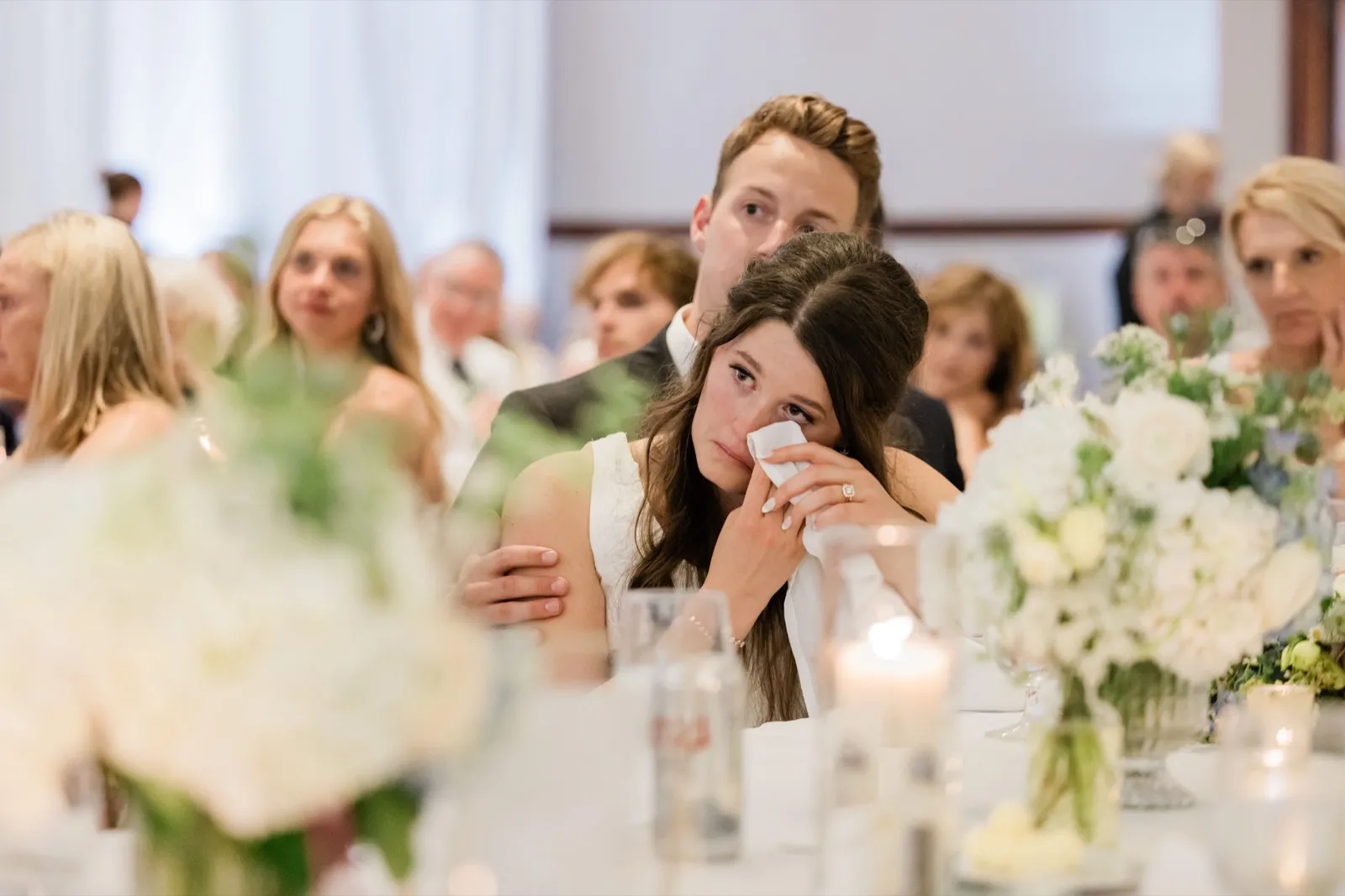 The bride wipes away a tear with a tissue while the groom holds her from behind during the toasts, framed by soft foreground florals — Tim Larsen Photography, Brainerd Lakes MN