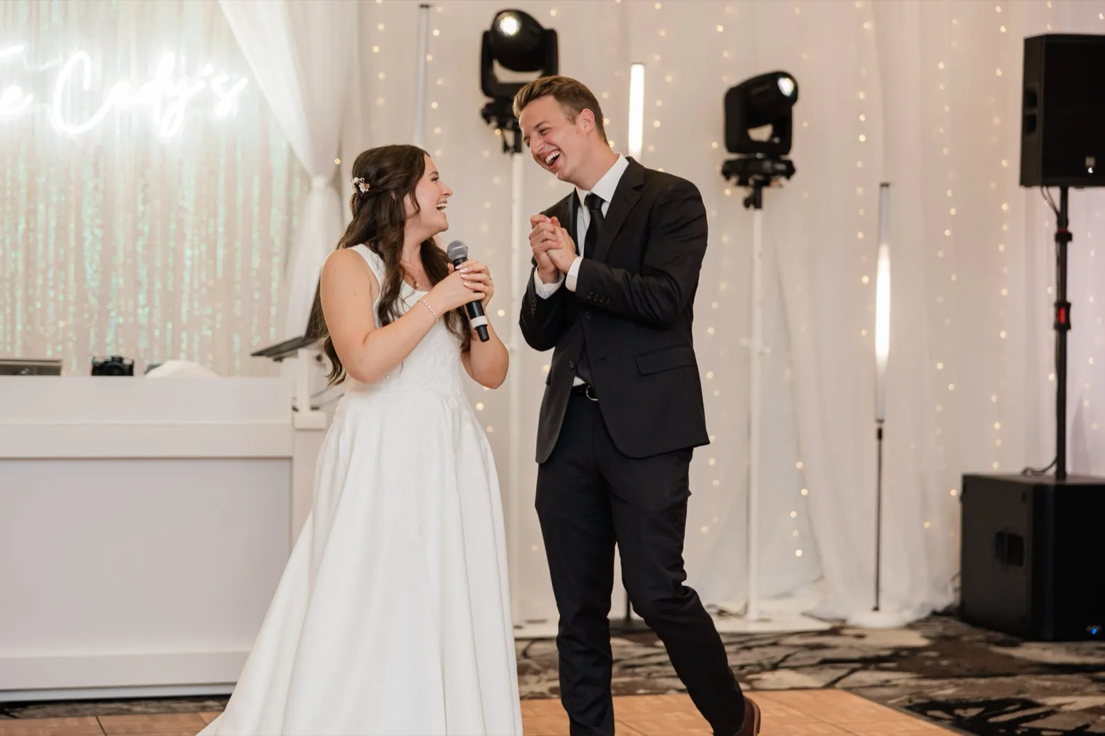 The bride speaks into a microphone while the groom laughs beside her in front of their neon sign backdrop — Tim Larsen Photography, Brainerd Lakes MN