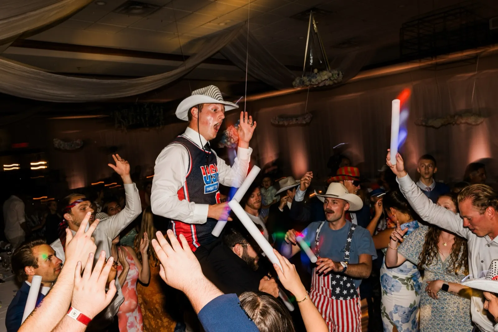 The groom is lifted on shoulders above a crowd of guests raising foam glow sticks under the draped ballroom ceiling — Tim Larsen Photography, Brainerd Lakes MN