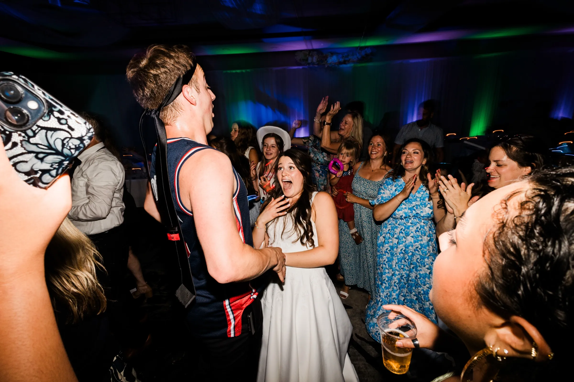 The bride presses a hand to her chest and reacts with delight toward the groom in a basketball jersey, guests cheering around them under blue light — Tim Larsen Photography, Brainerd Lakes MN