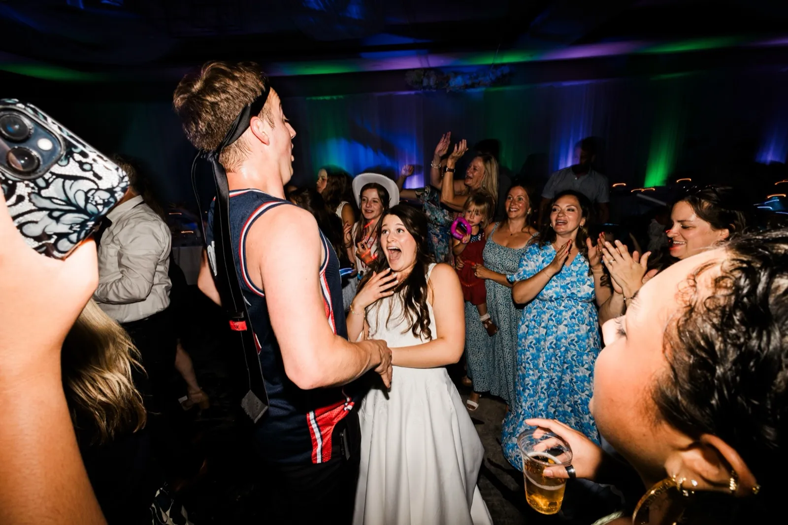 The bride presses a hand to her chest and reacts with delight toward the groom in a basketball jersey, guests cheering around them under blue light — Tim Larsen Photography, Brainerd Lakes MN