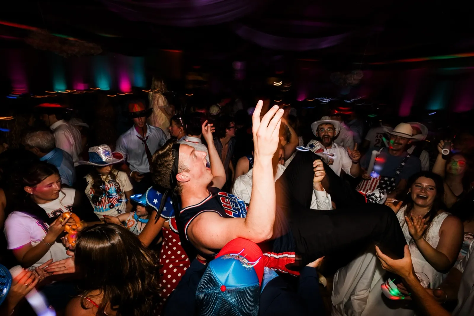 A guest in a USA tank top is lifted horizontally above the packed dance floor while others reach up to hold him — Tim Larsen Photography, Brainerd Lakes MN