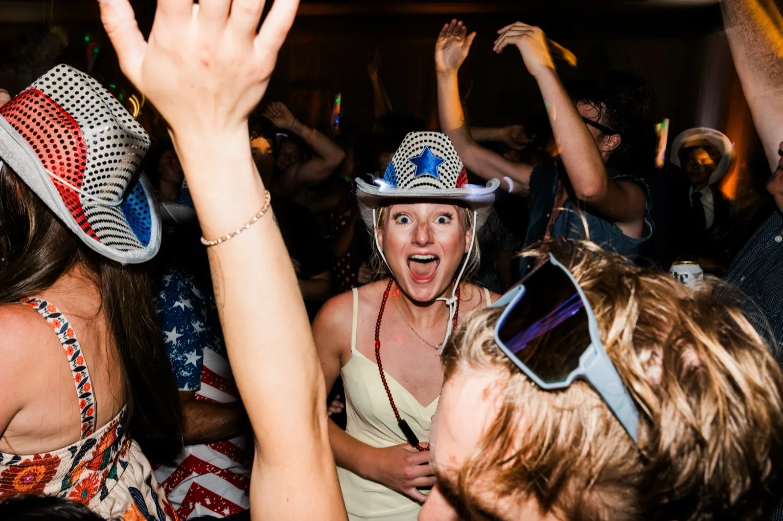 A guest in a star-spangled cowboy hat throws her mouth wide open in delight amid a crowd of raised arms — Tim Larsen Photography, Brainerd Lakes MN