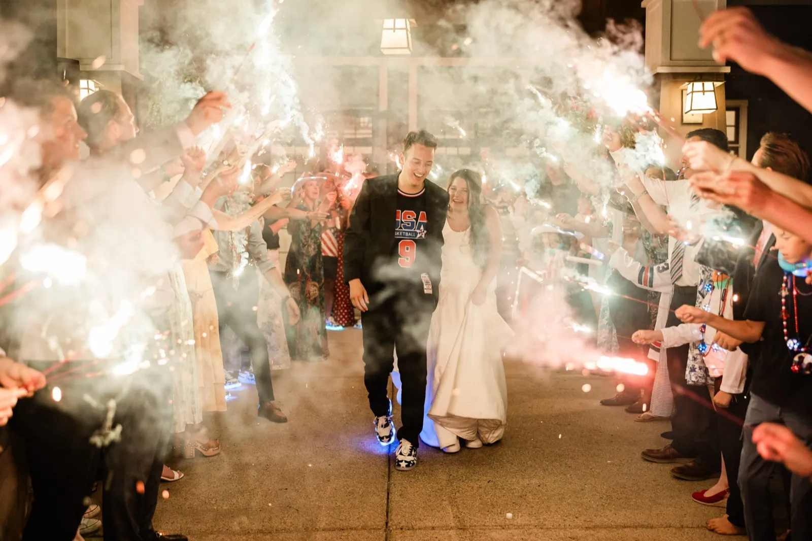 The bride and groom walk toward the camera through a tunnel of sparklers and smoke as guests line both sides — Tim Larsen Photography, Brainerd Lakes MN