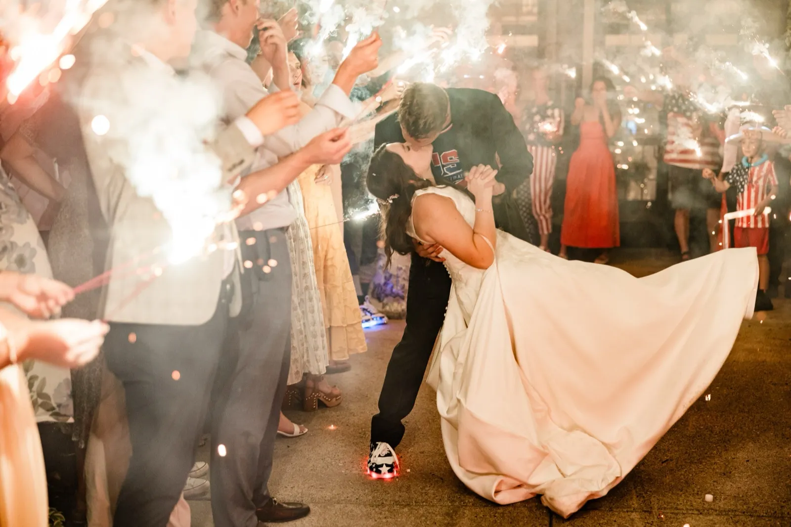 The groom dips the bride into a kiss, her gown sweeping across the pavement, framed by sparklers and smoke on both sides — Tim Larsen Photography, Brainerd Lakes MN