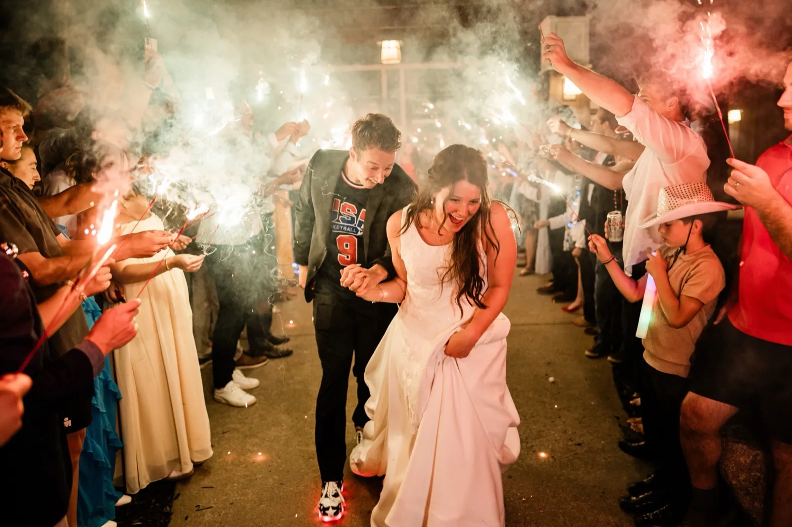 The bride and groom laugh hand in hand through a tunnel of sparklers at their Grand View Lodge send-off, the groom in a Team USA jersey — Tim Larsen Photography, Brainerd Lakes MN