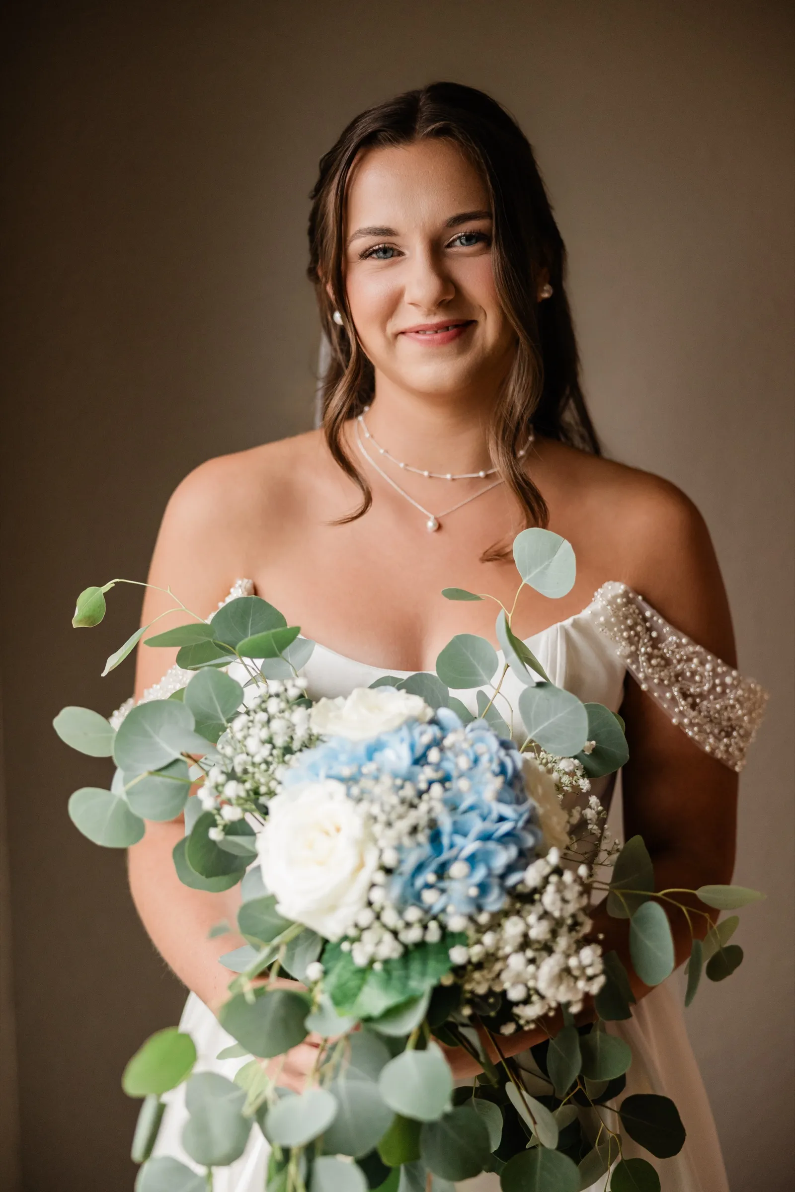 The bride holds her bouquet up toward the camera in window light, the flowers sharper than her quiet expression behind them — Tim Larsen Photography, Brainerd Lakes MN