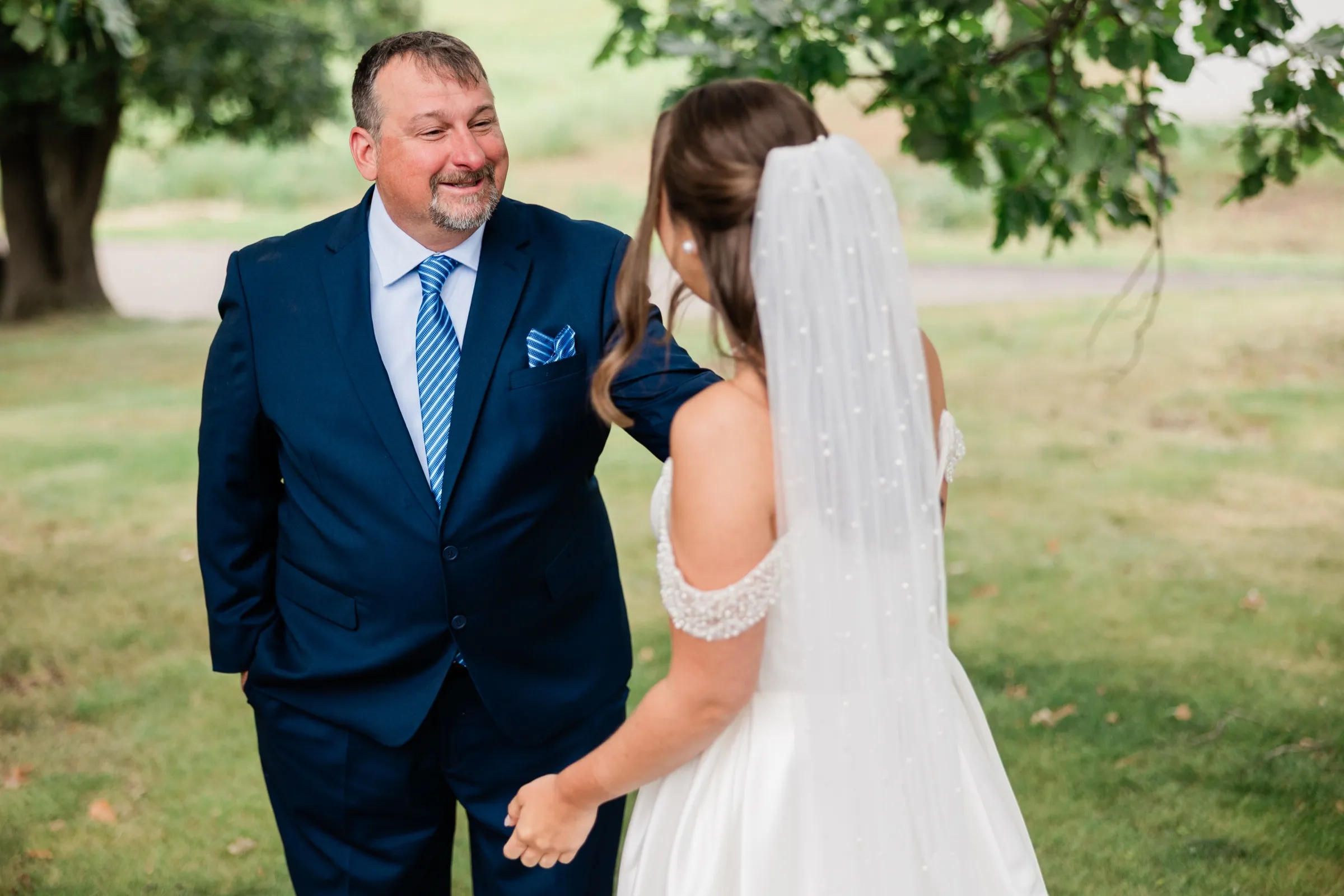 The bride's father looks at her with a closed-lipped smile on a shaded lawn, the bride's pearl veil filling the right side of the frame — Tim Larsen Photography, Brainerd Lakes MN