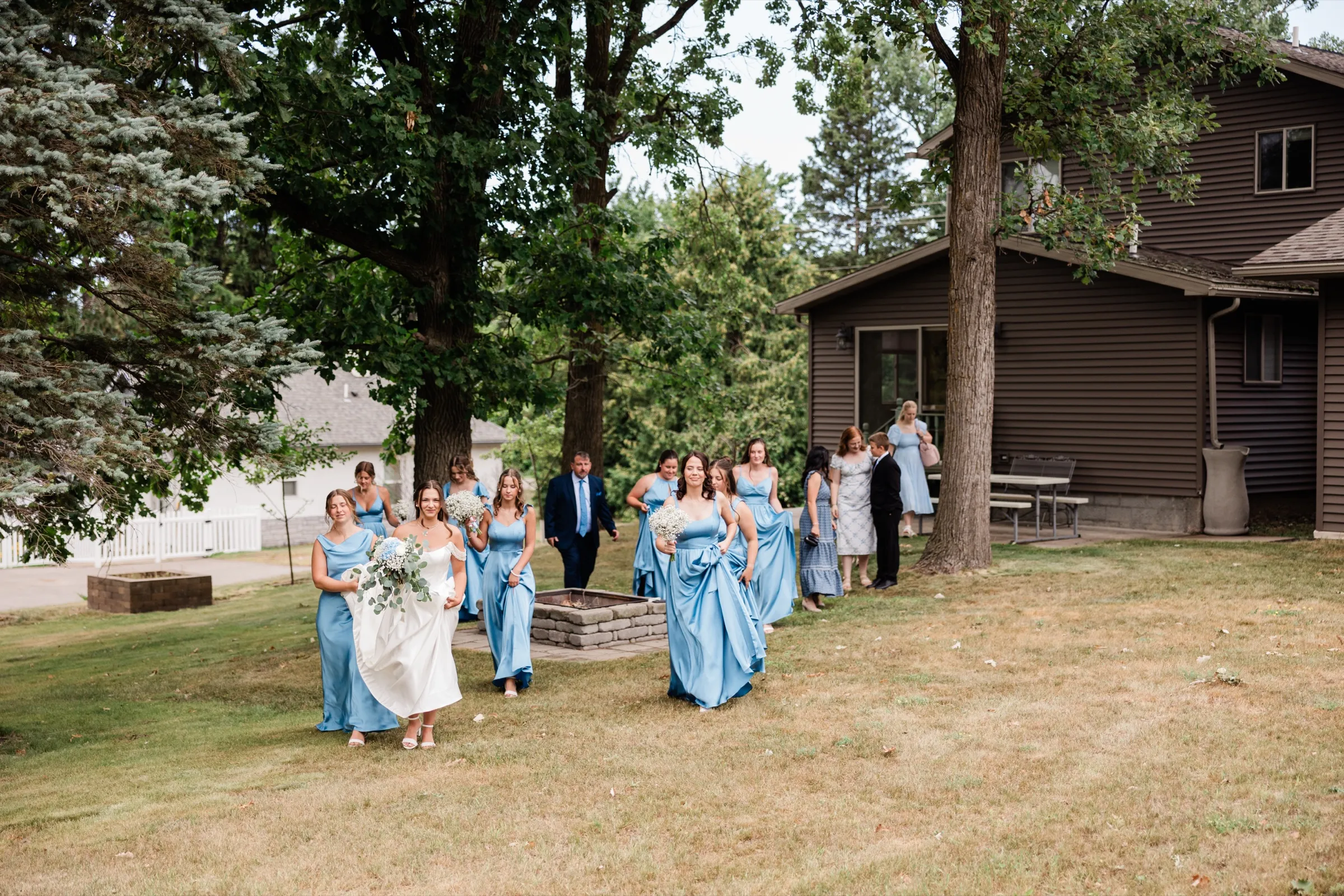 The bride and her bridesmaids in blue walk across her grandpa's wide backyard, the house shaded by oak trees behind them — Tim Larsen Photography, Brainerd Lakes MN