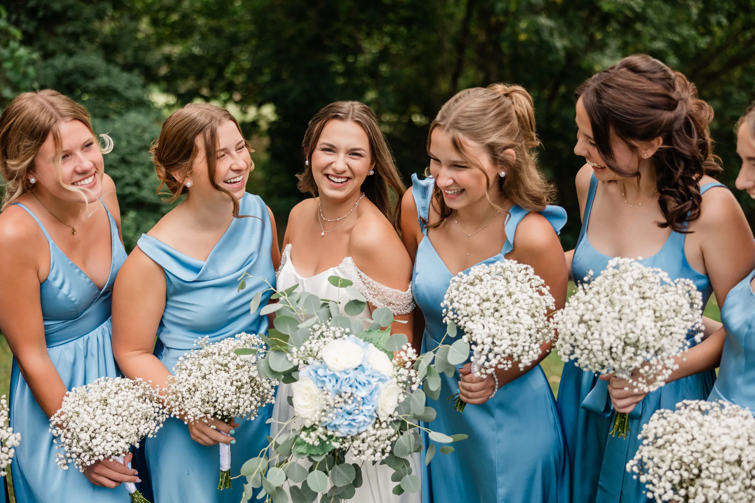 Bridesmaids in blue lean in laughing on either side of the bride, their baby's breath bouquets filling the lower frame — Tim Larsen Photography, Brainerd Lakes MN