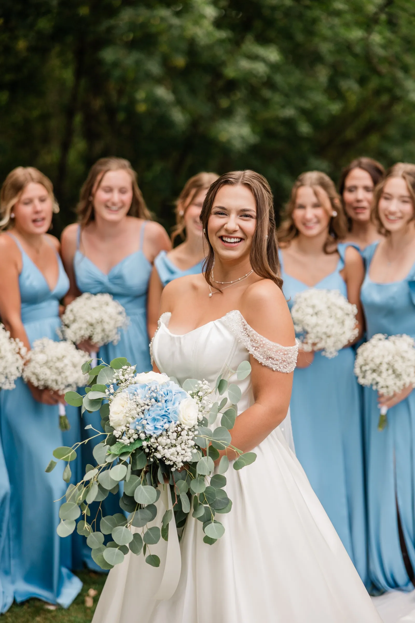 The bride smiles in sharp focus holding her bouquet while her bridesmaids blur into the background behind her — Tim Larsen Photography, Brainerd Lakes MN