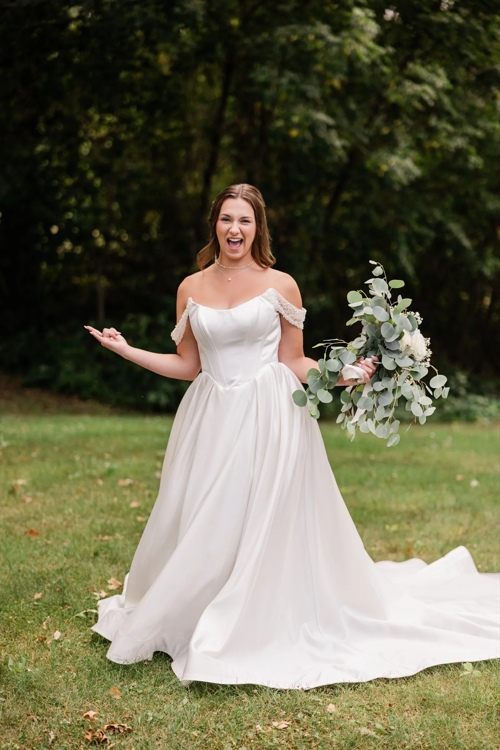 The bride laughs with her arms open, holding her bouquet, standing on a shaded lawn with the train of her gown fanned out behind her — Tim Larsen Photography, Brainerd Lakes MN
