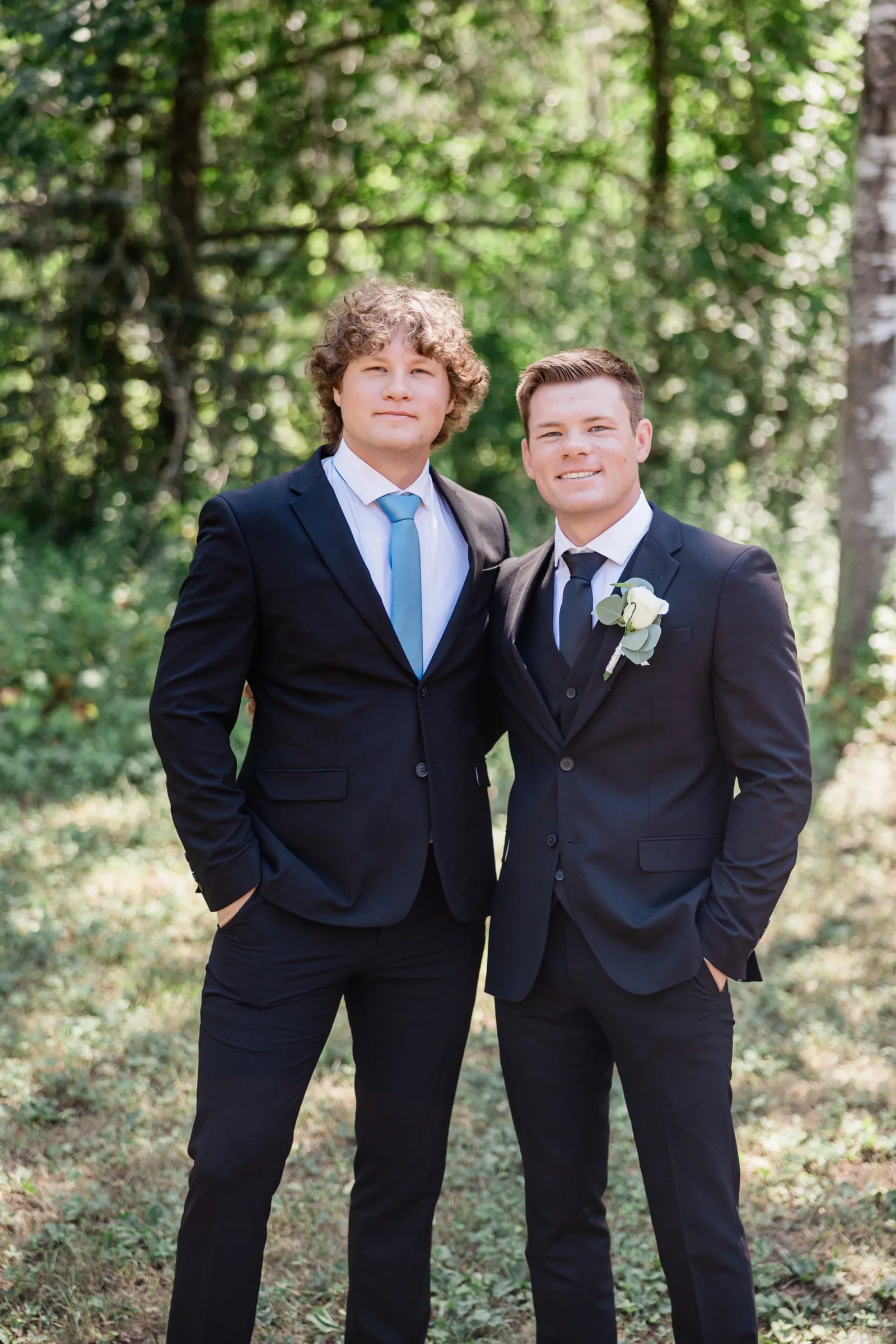 The groom and a groomsman stand arm in arm in a grassy clearing at the edge of the woods, both in navy suits — Tim Larsen Photography, Brainerd Lakes MN