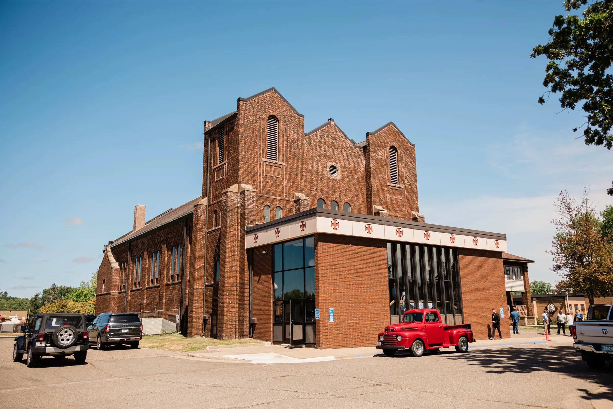 A red brick Catholic church with tall arched windows stands against a clear blue sky, a vintage red pickup truck parked at its entrance — Tim Larsen Photography, Brainerd Lakes MN