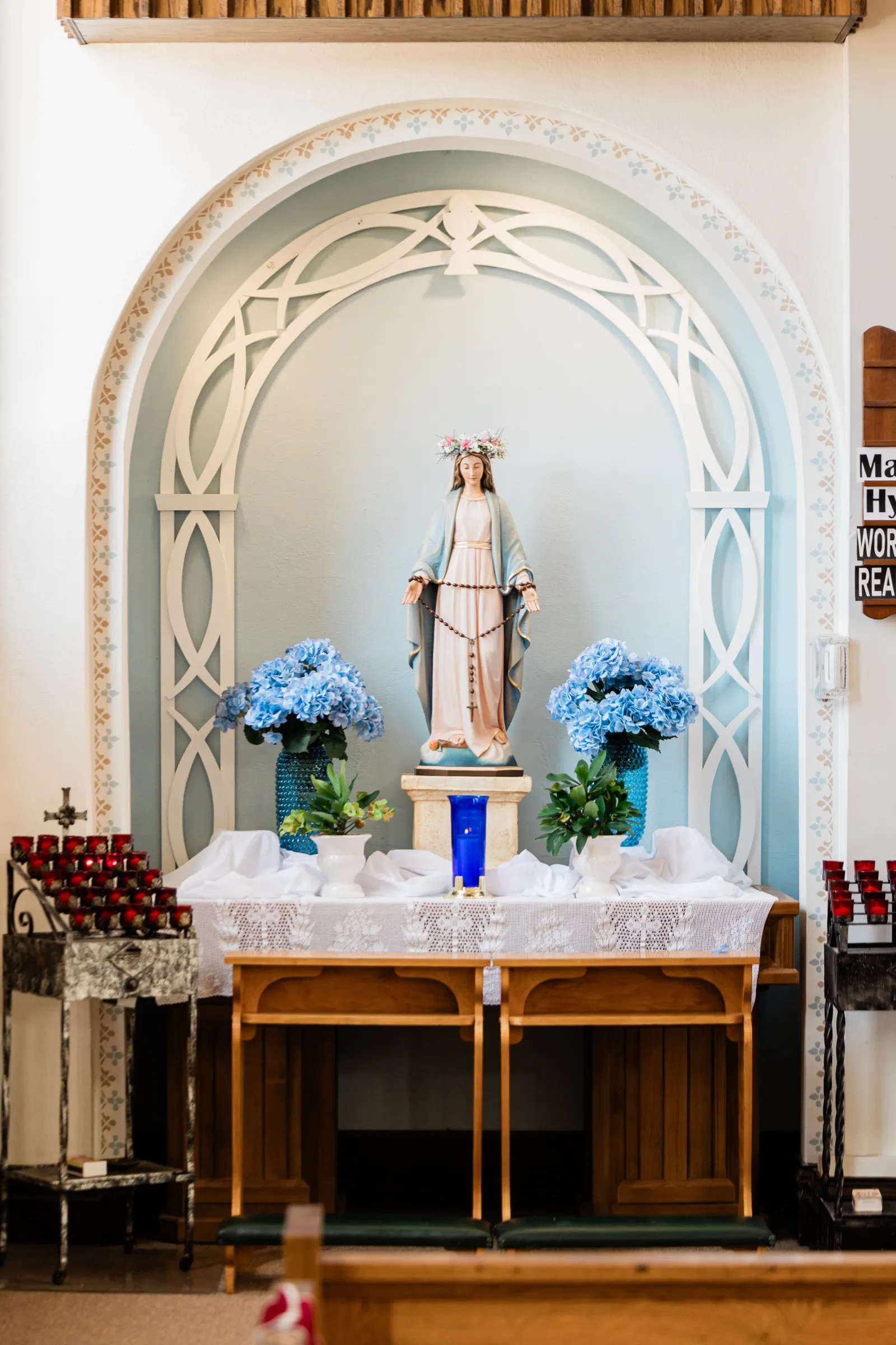 A statue of Mary stands in a pale blue arched alcove flanked by two vases of blue hydrangea on a lace-covered table — Tim Larsen Photography, Brainerd Lakes MN