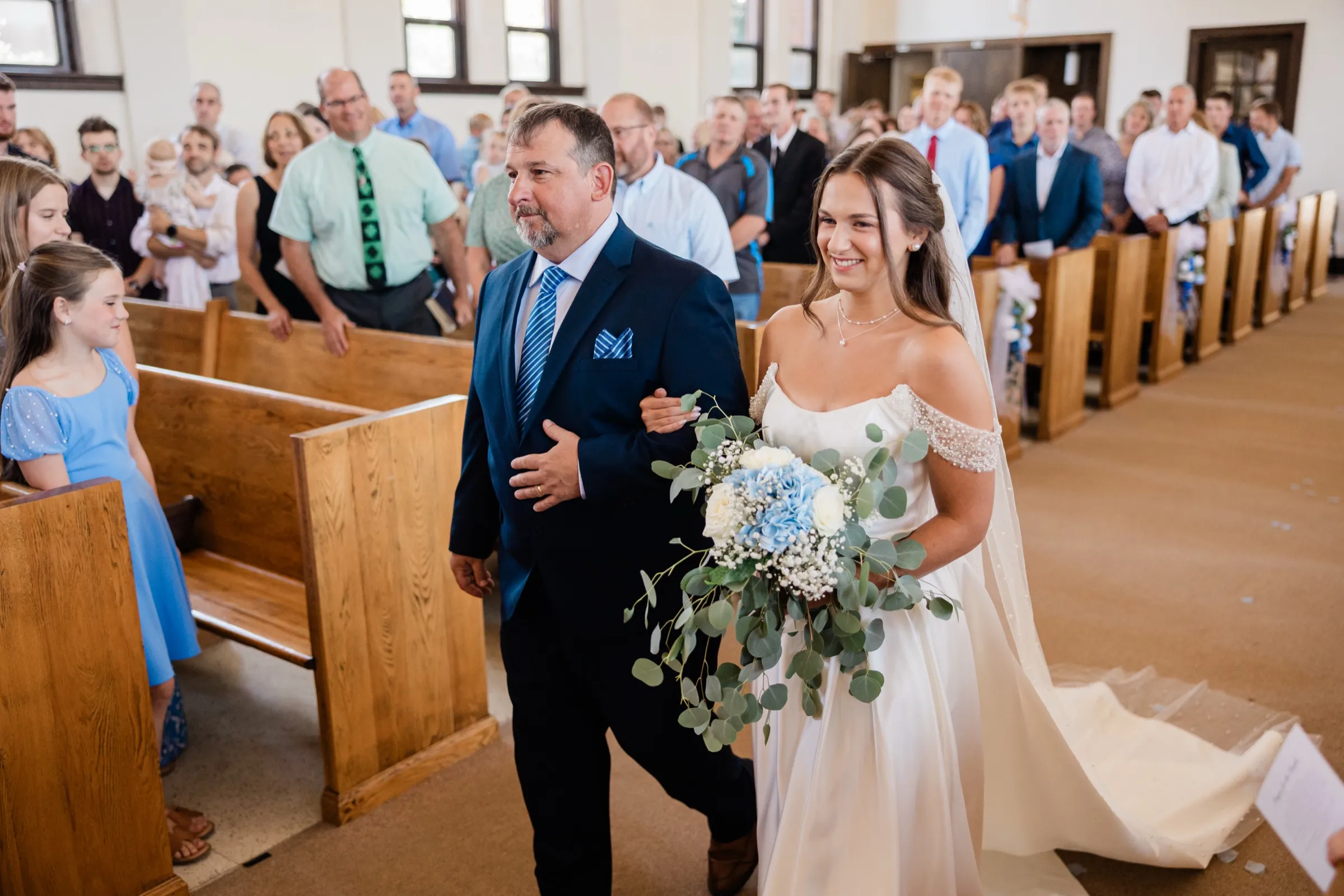 The bride walks up the aisle on her father's arm, smiling, holding her blue hydrangea bouquet as guests stand behind her — Tim Larsen Photography, Brainerd Lakes MN