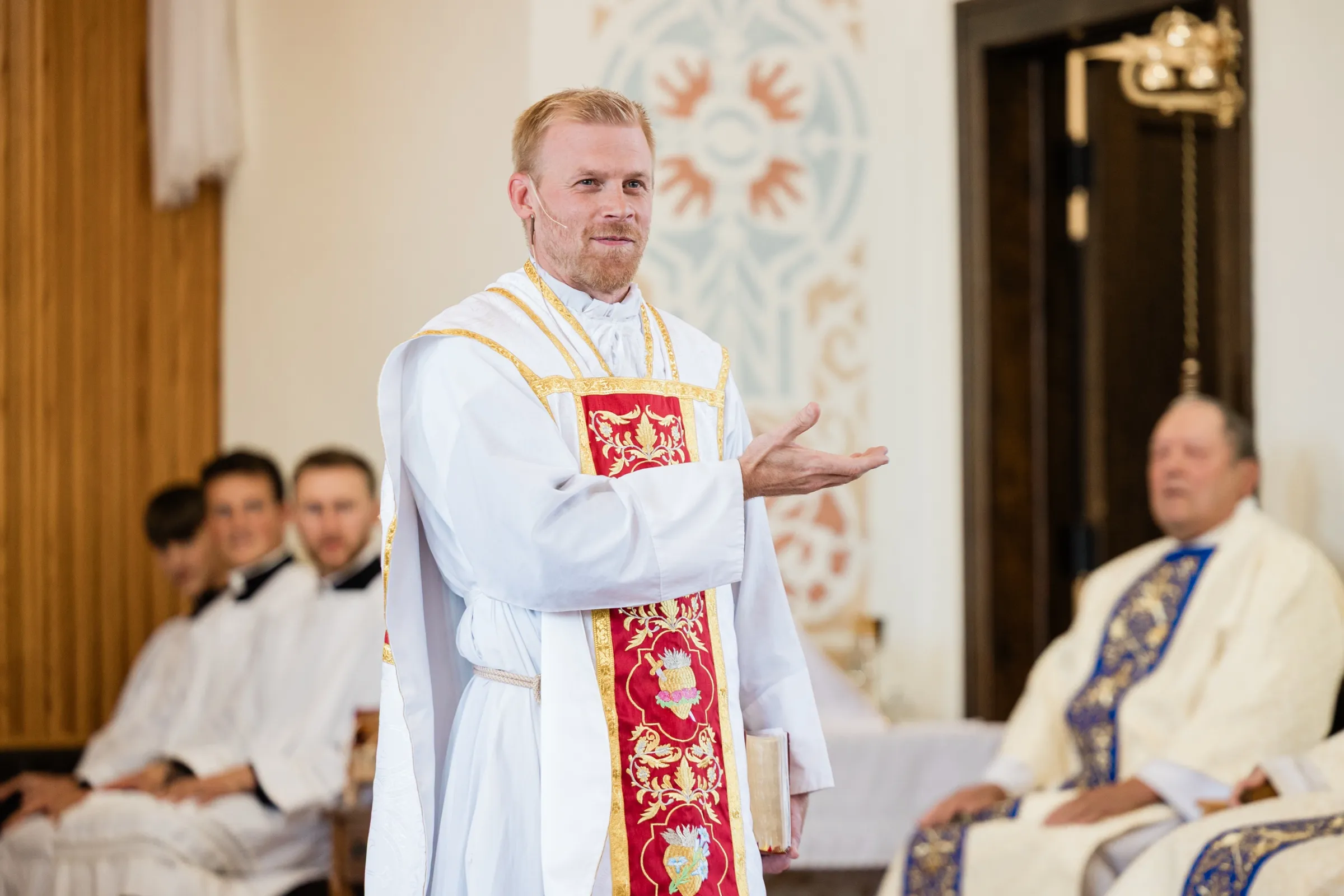 A priest in white and red vestments gestures with one hand while speaking during the wedding Mass, with a seated concelebrant and altar servers behind him — Tim Larsen Photography, Brainerd Lakes MN