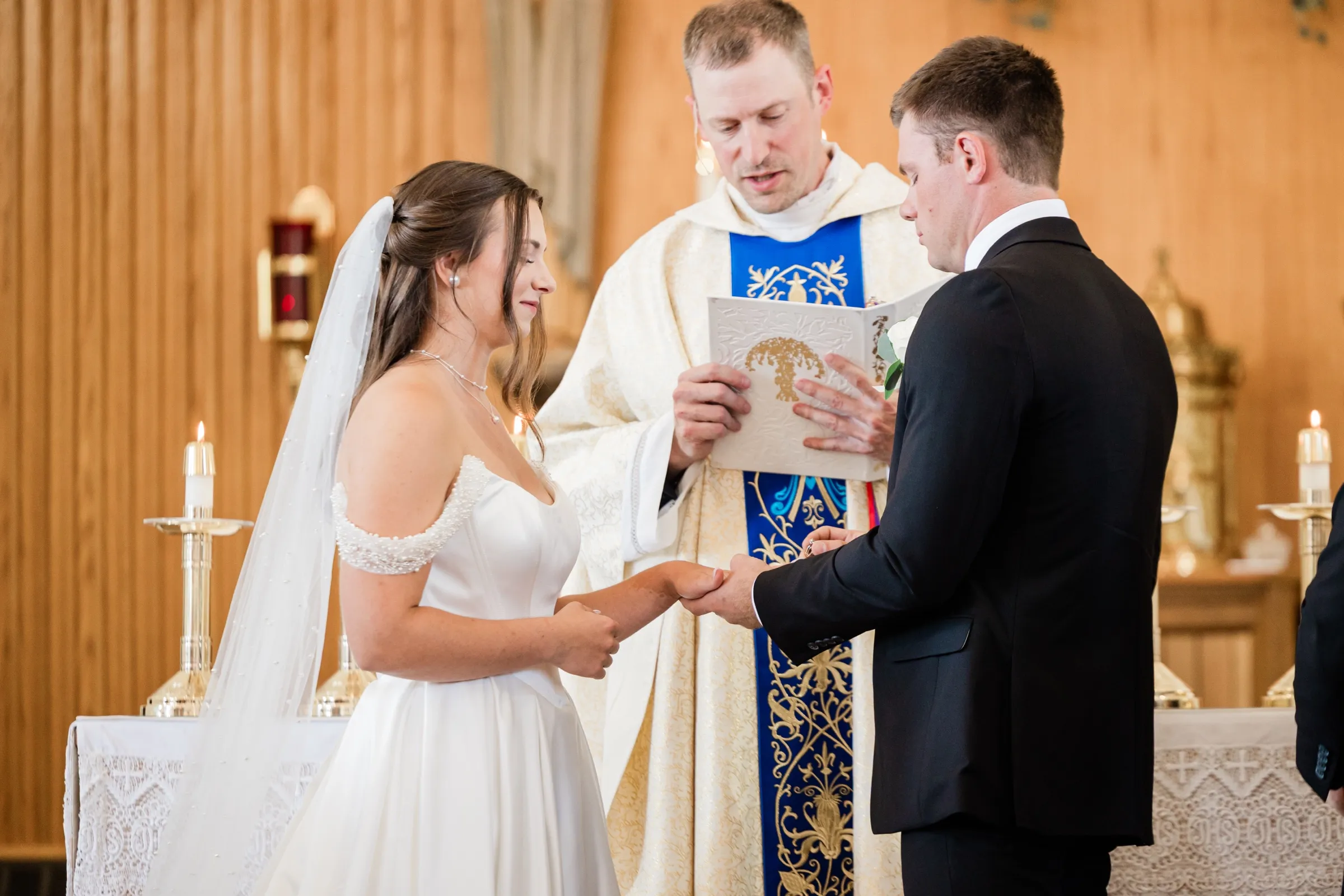 The groom slides a ring onto the bride's finger as the priest reads from a book held open between them — Tim Larsen Photography, Brainerd Lakes MN