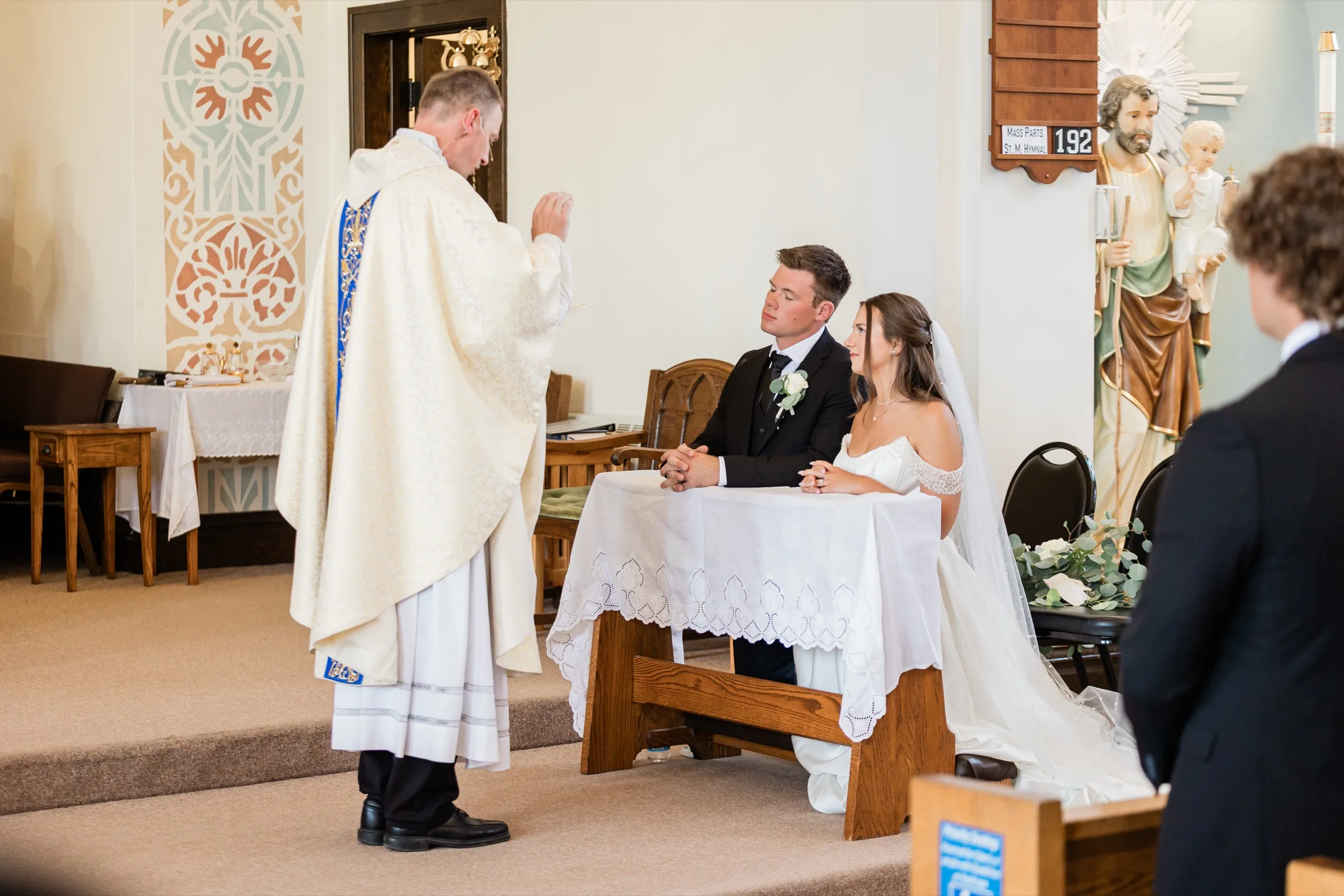 The bride and groom kneel side by side at a linen-covered prayer bench with hands folded as the priest raises his hand in blessing — Tim Larsen Photography, Brainerd Lakes MN