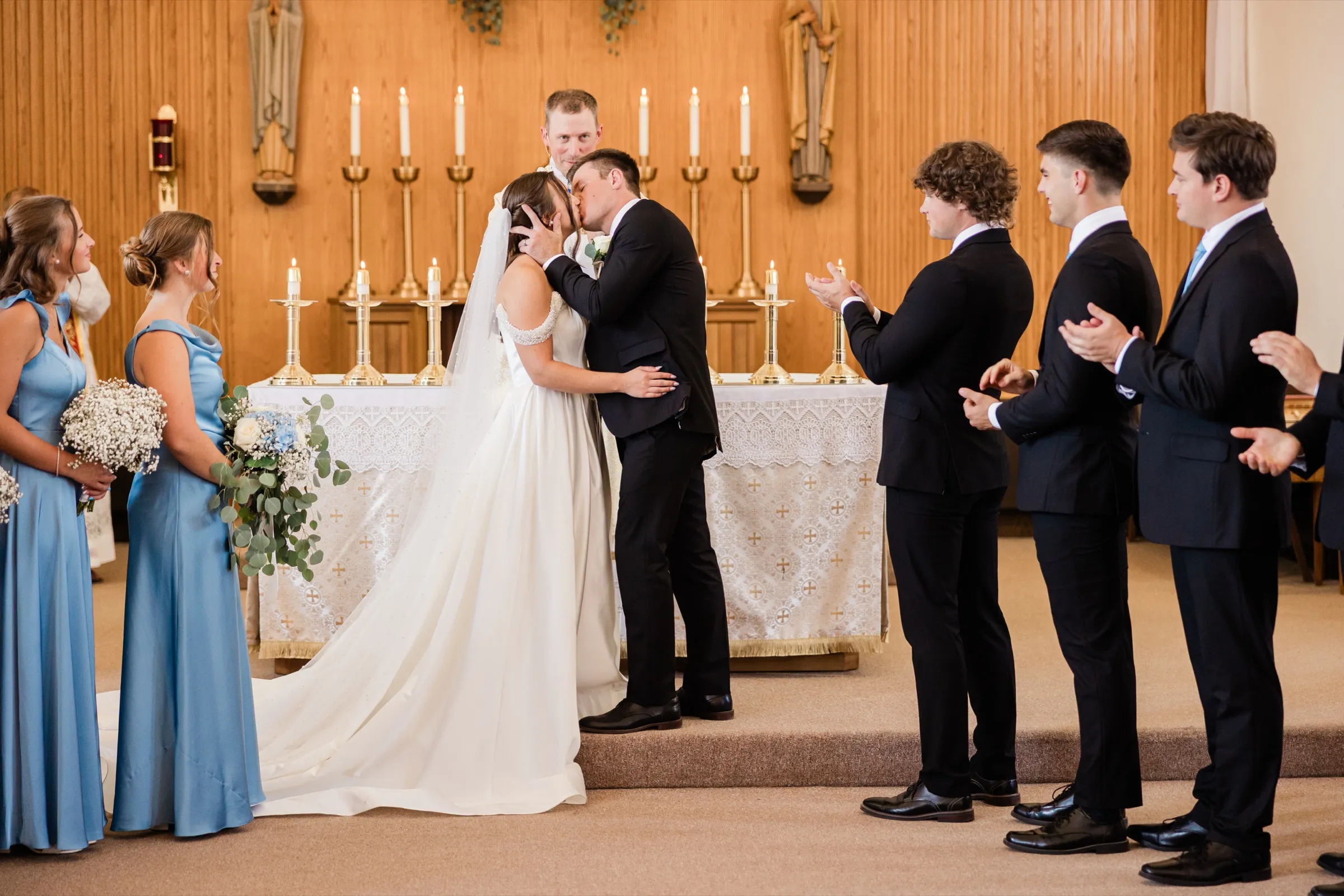 The groom holds the bride's face as they kiss in front of the candlelit altar while groomsmen applaud to the right and bridesmaids watch from the left — Tim Larsen Photography, Brainerd Lakes MN