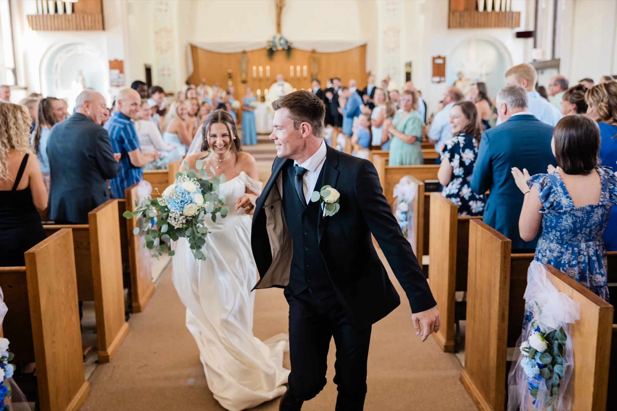 The groom walks quickly toward the camera with a wide smile, jacket open, as the bride laughs behind him with her bouquet and the applauding congregation fills both sides of the aisle — Tim Larsen Photography, Brainerd Lakes MN