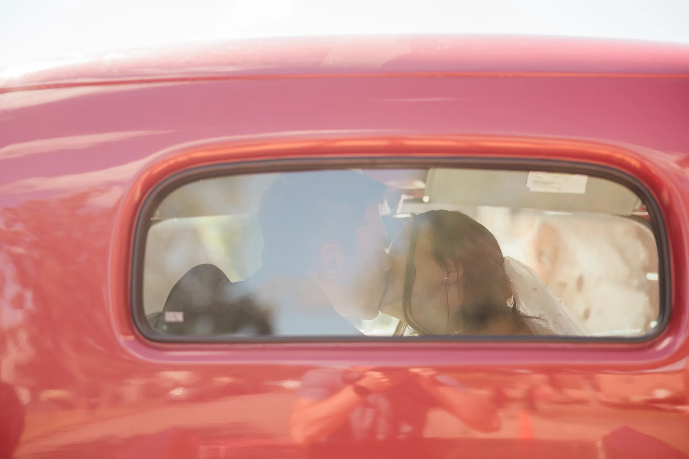 The bride and groom kiss inside the cab of a red pickup, seen in silhouette through the dusty rear window surrounded by red painted metal — Tim Larsen Photography, Brainerd Lakes MN