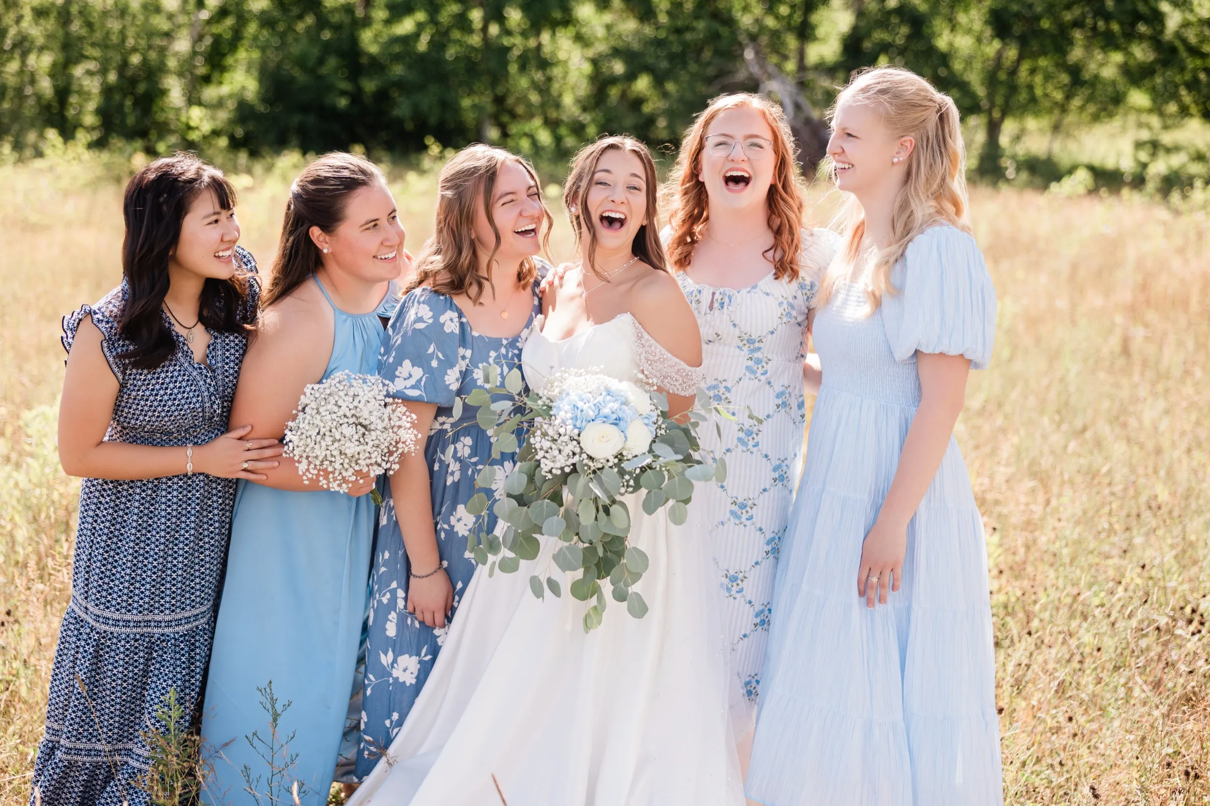 The bride and five friends in blue dresses laugh with their heads thrown back in a sunlit field, arms wrapped around each other — Tim Larsen Photography, Brainerd Lakes MN
