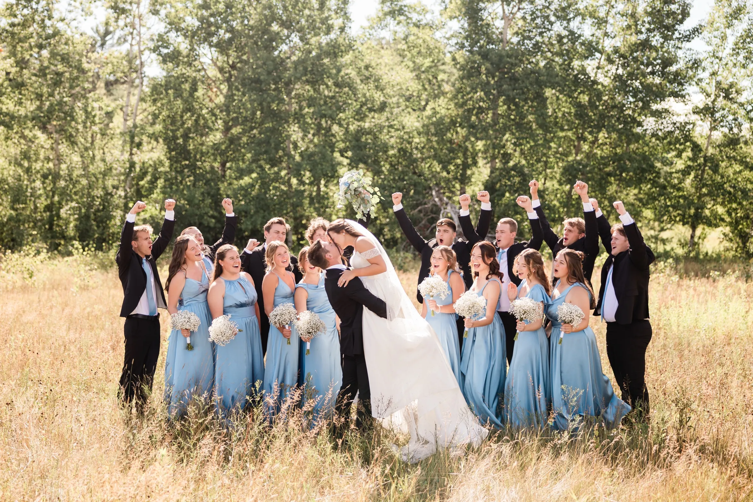 The groom dips the bride into a kiss in a dry field while the wedding party around them shout and throw their fists and bouquets into the air — Tim Larsen Photography, Brainerd Lakes MN