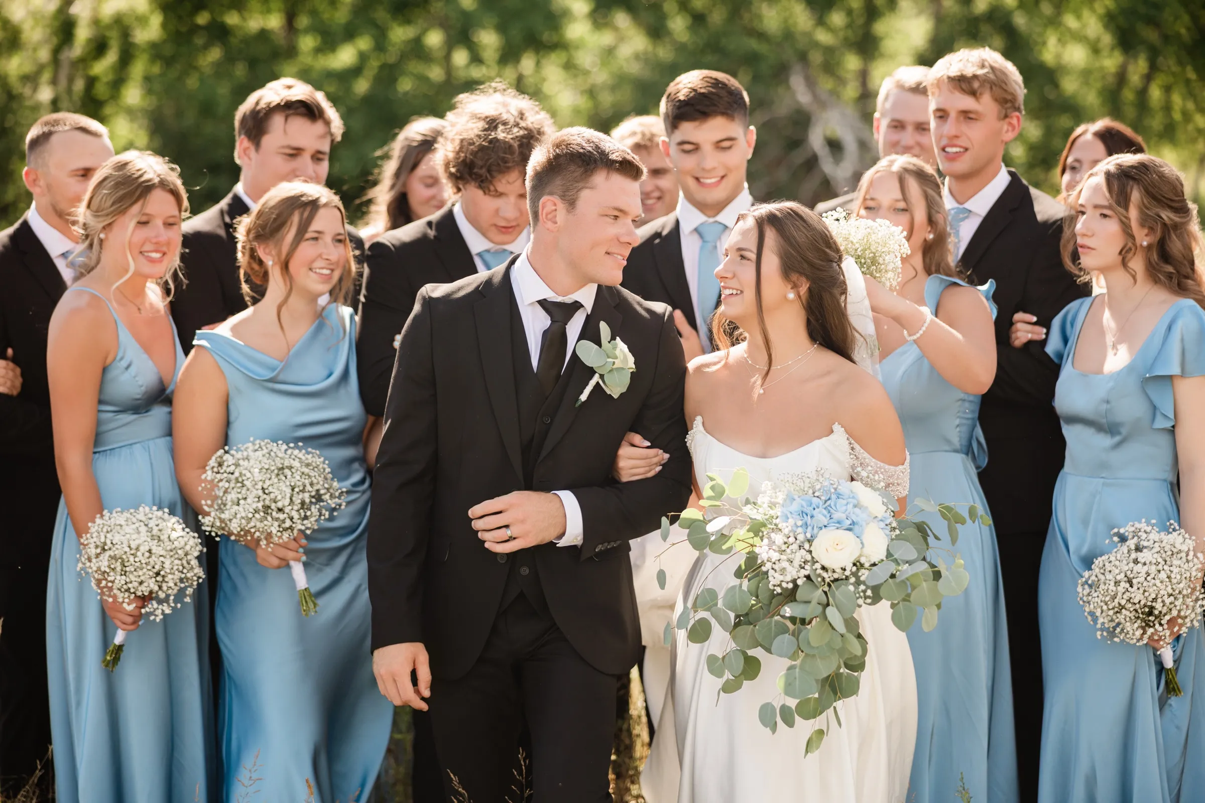 The bride looks up at the groom with a smile as they walk arm in arm through a field, the wedding party blurred behind them — Tim Larsen Photography, Brainerd Lakes MN