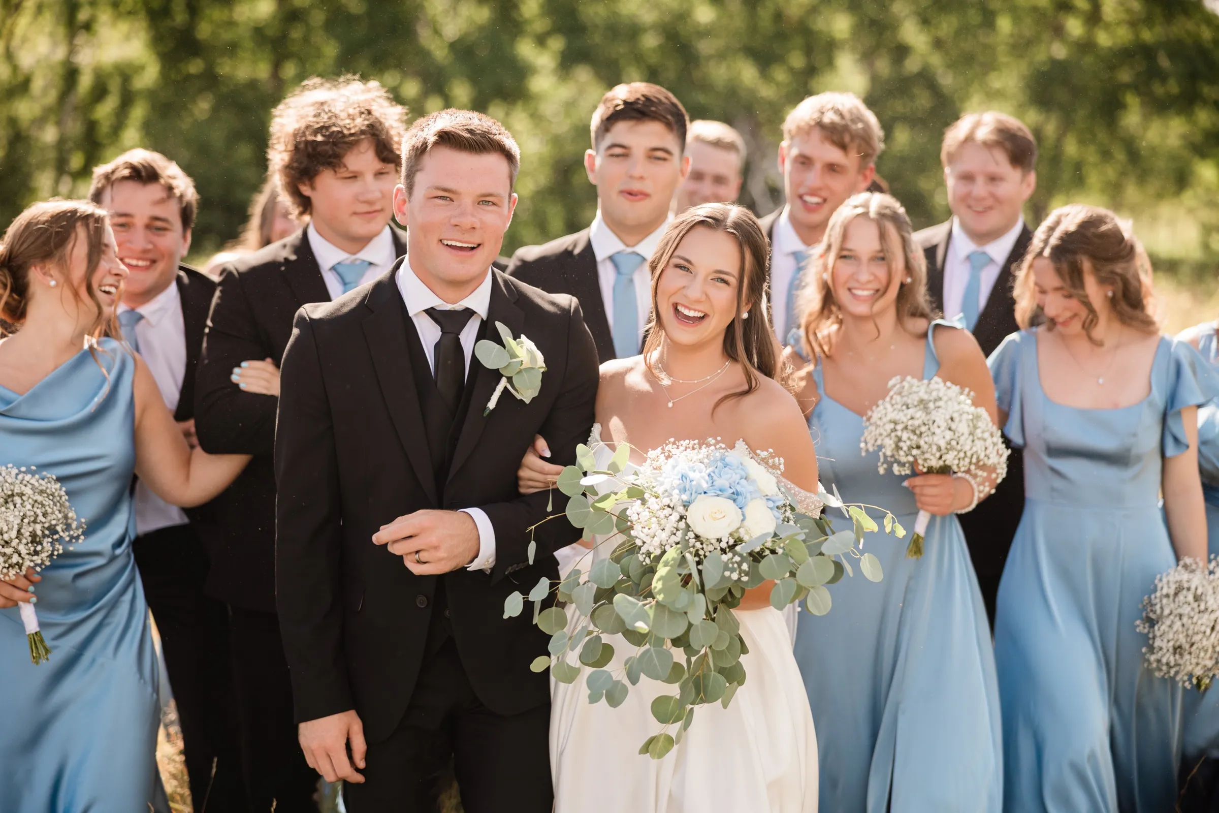 The bride and groom walk toward the camera laughing together, arms linked, with the wedding party following in a loose crowd behind them — Tim Larsen Photography, Brainerd Lakes MN