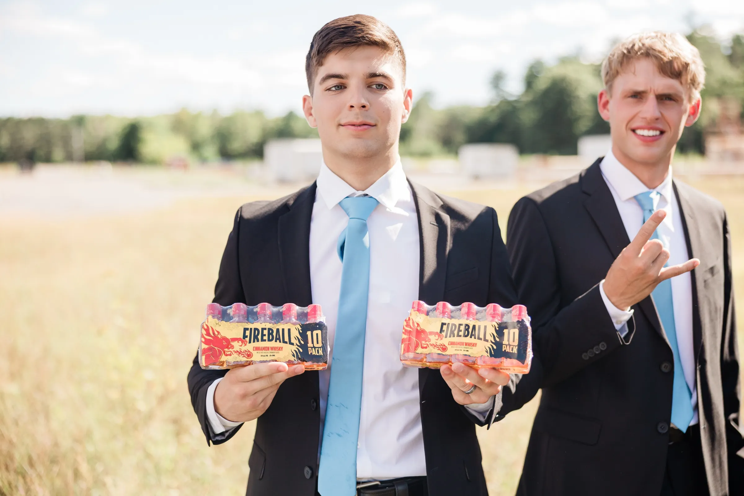 Two groomsmen in black suits and light blue ties hold up ten-packs of cinnamon whisky shooters toward the camera on the open grounds of the fairgrounds — Tim Larsen Photography, Brainerd Lakes MN