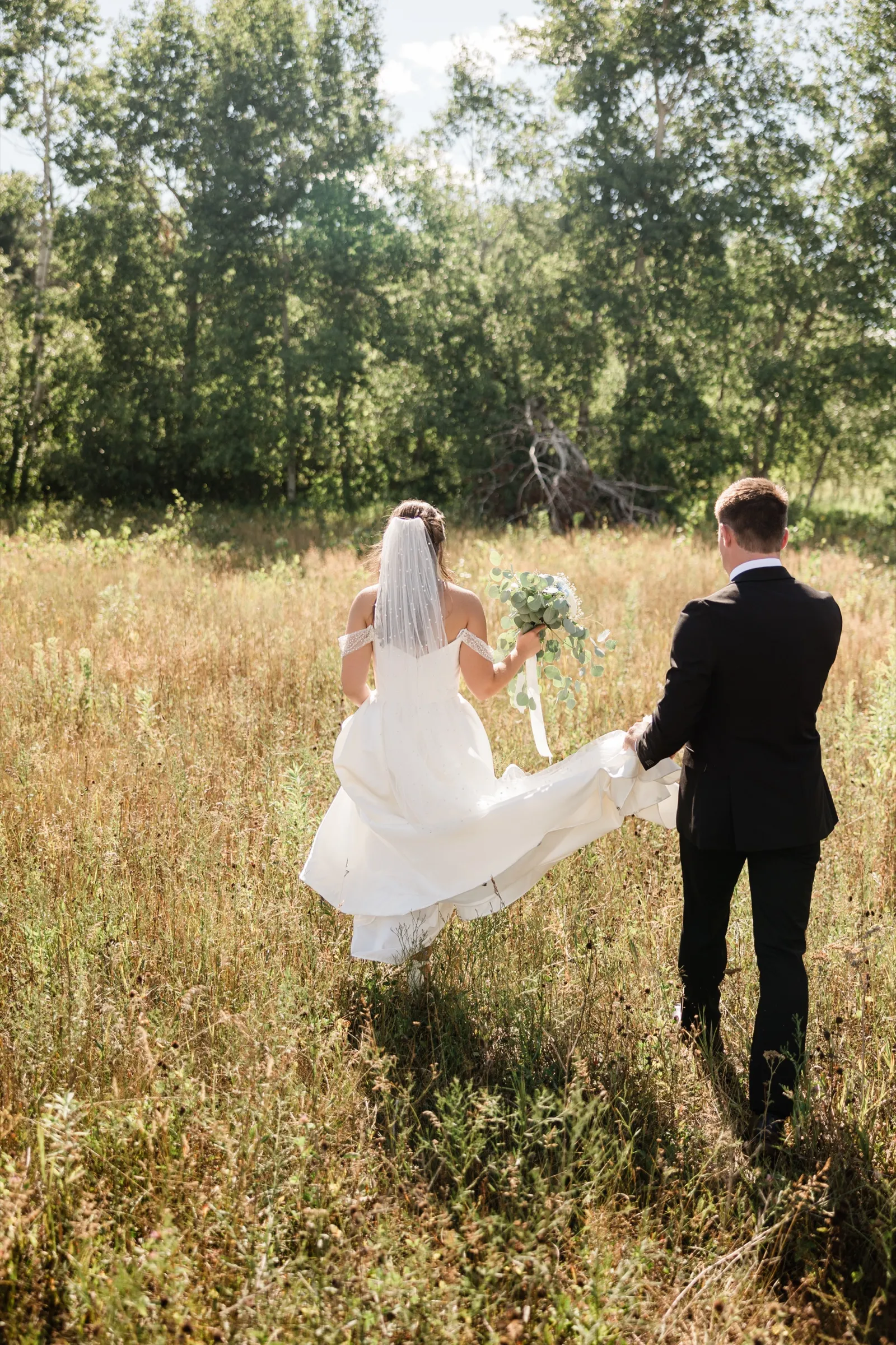 Seen from behind, the bride walks into a field of tall dry grass with her bouquet at her side while the groom walks beside her carrying the trailing end of her dress — Tim Larsen Photography, Brainerd Lakes MN